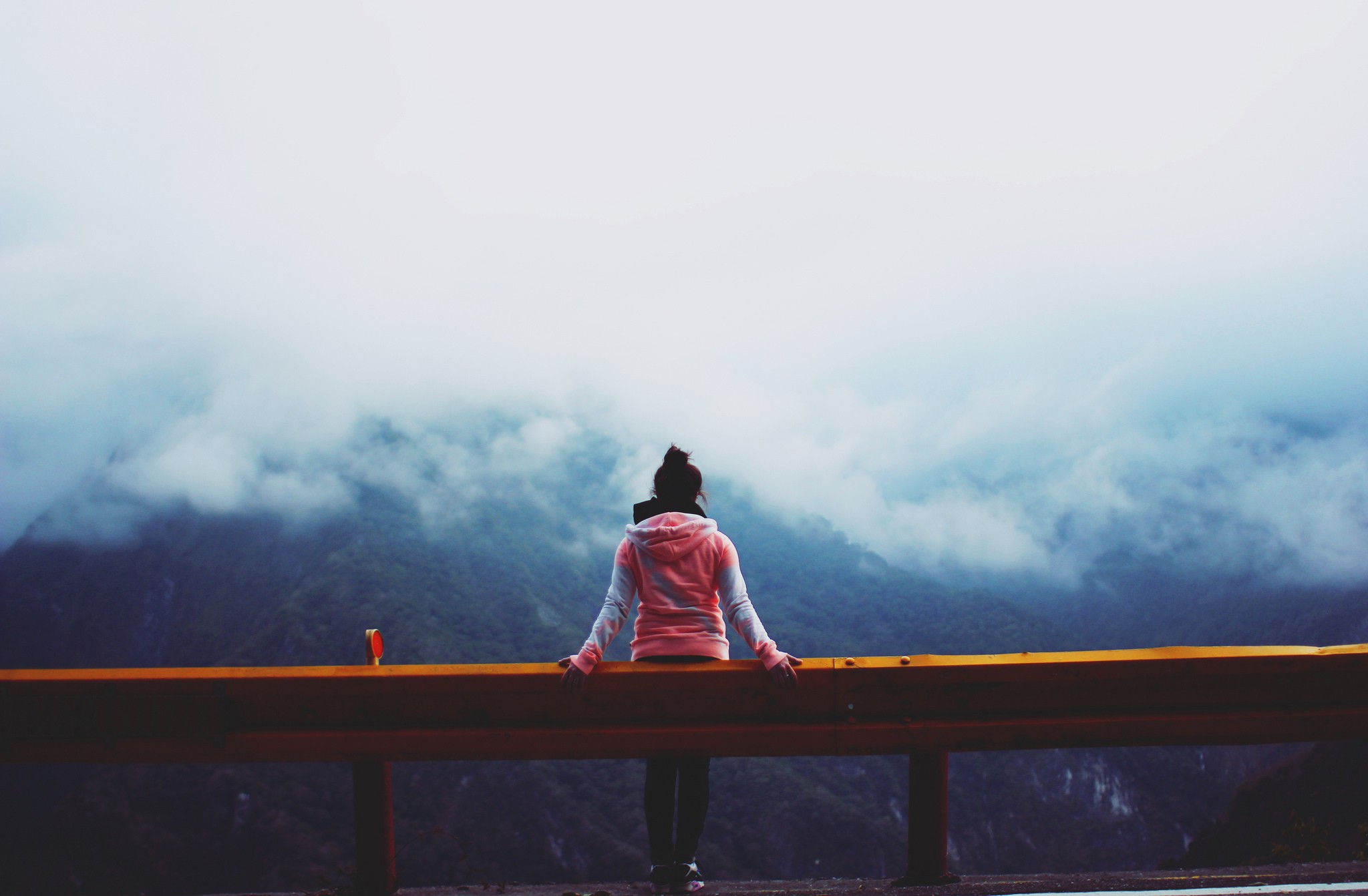 Woman in hoodie sitting on guardrail overlooking foggy mountains HD Wallpaper