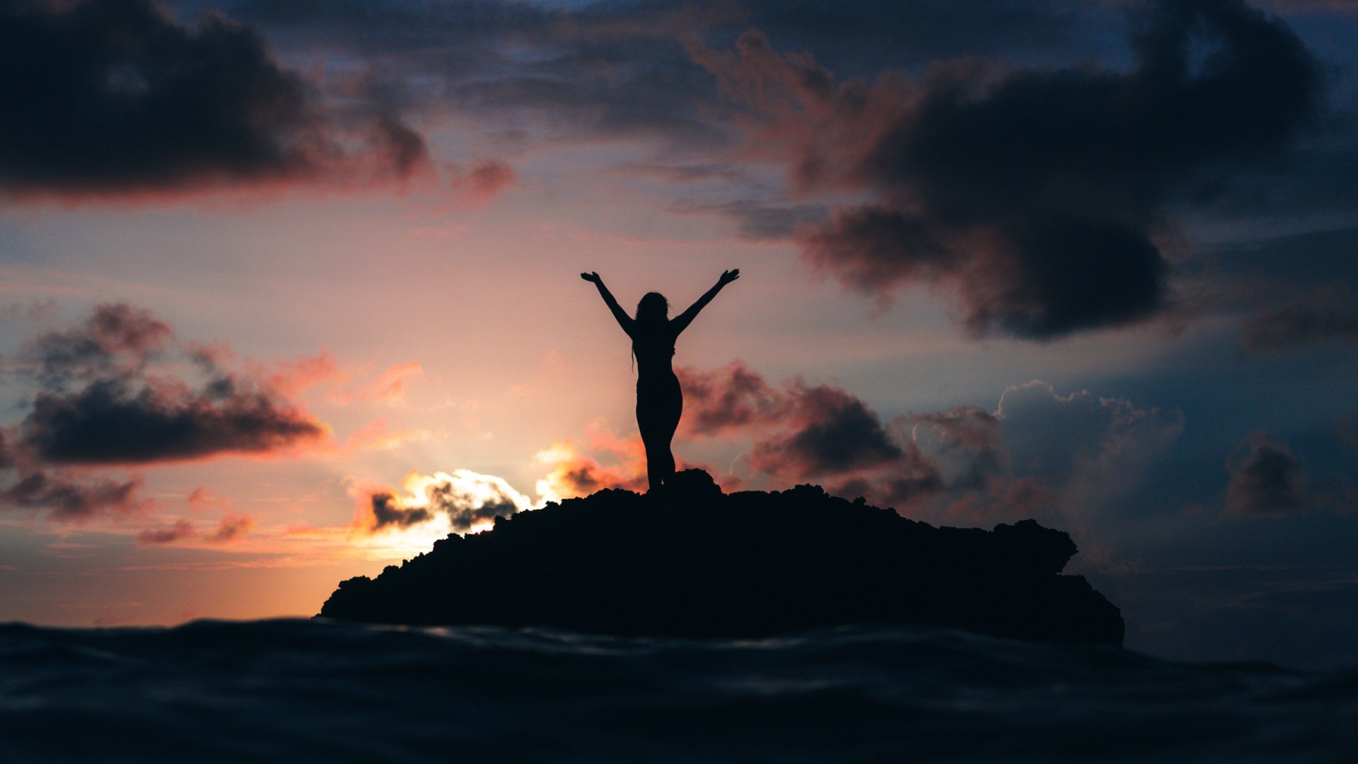 Silhouette of a woman with arms raised on a rock at sunset HD Wallpaper