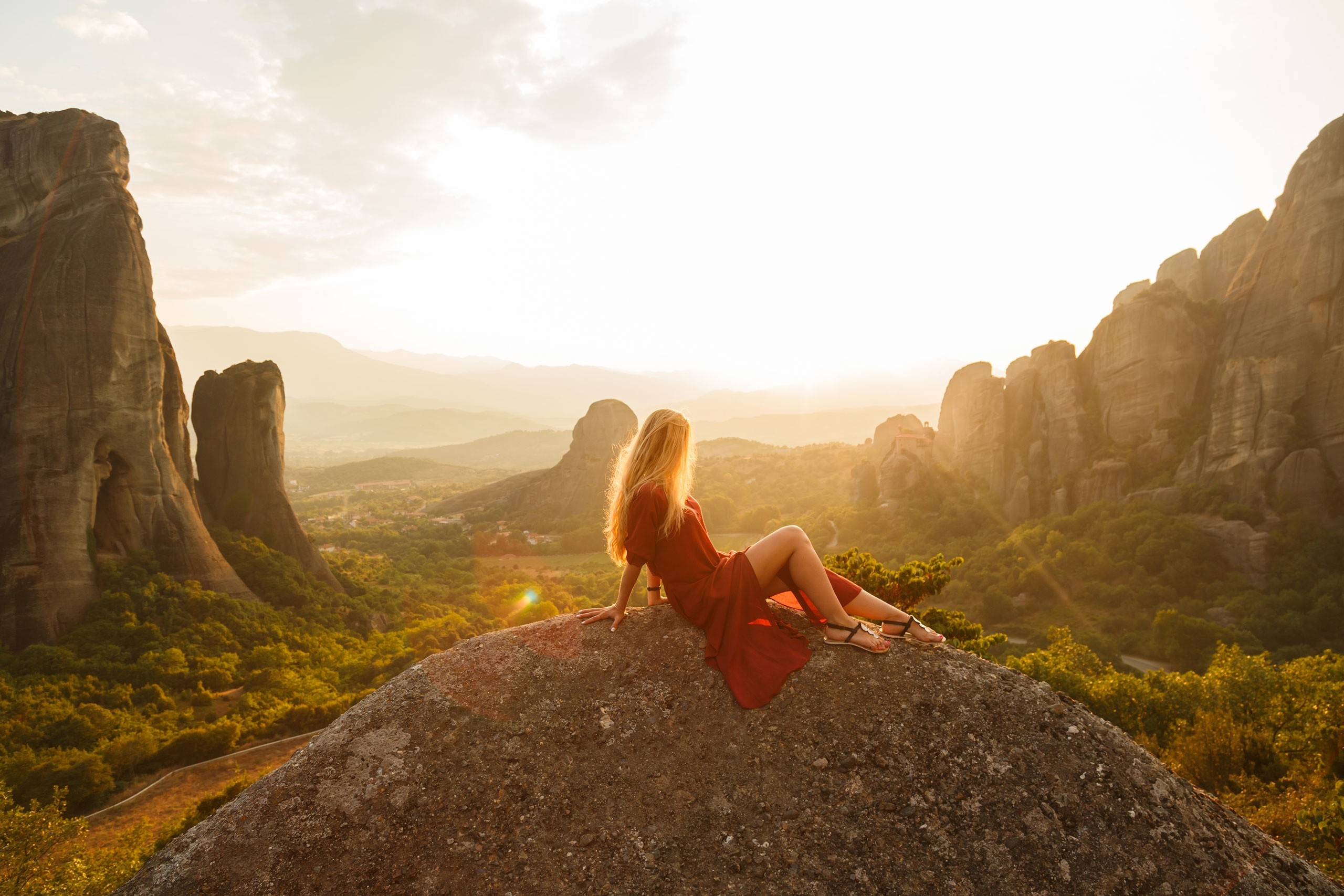 Woman in red dress sitting on rock overlooking Meteora landscape at sunset HD Wallpaper