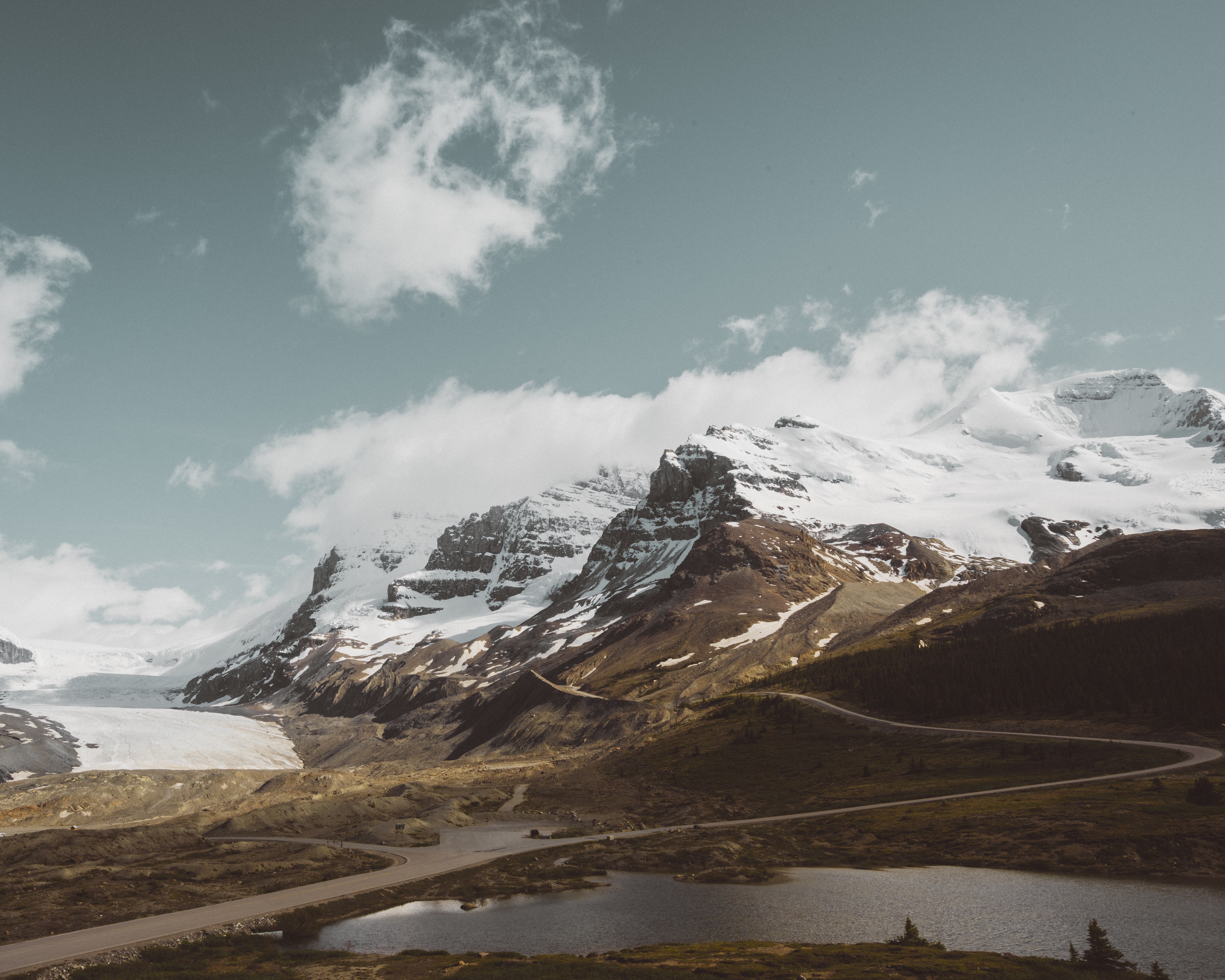 Snow-capped mountains and a winding road near a lake HD Wallpaper