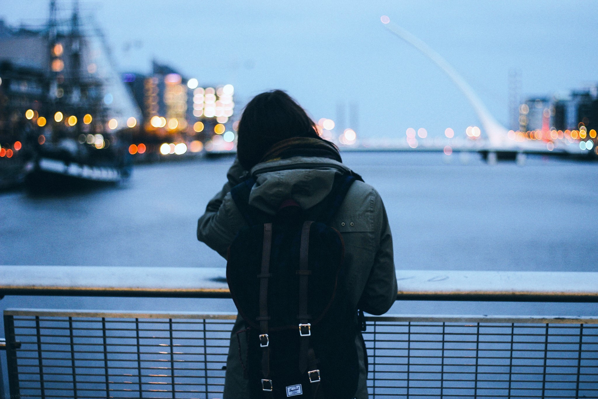 Woman with backpack looking at city lights across river HD Wallpaper
