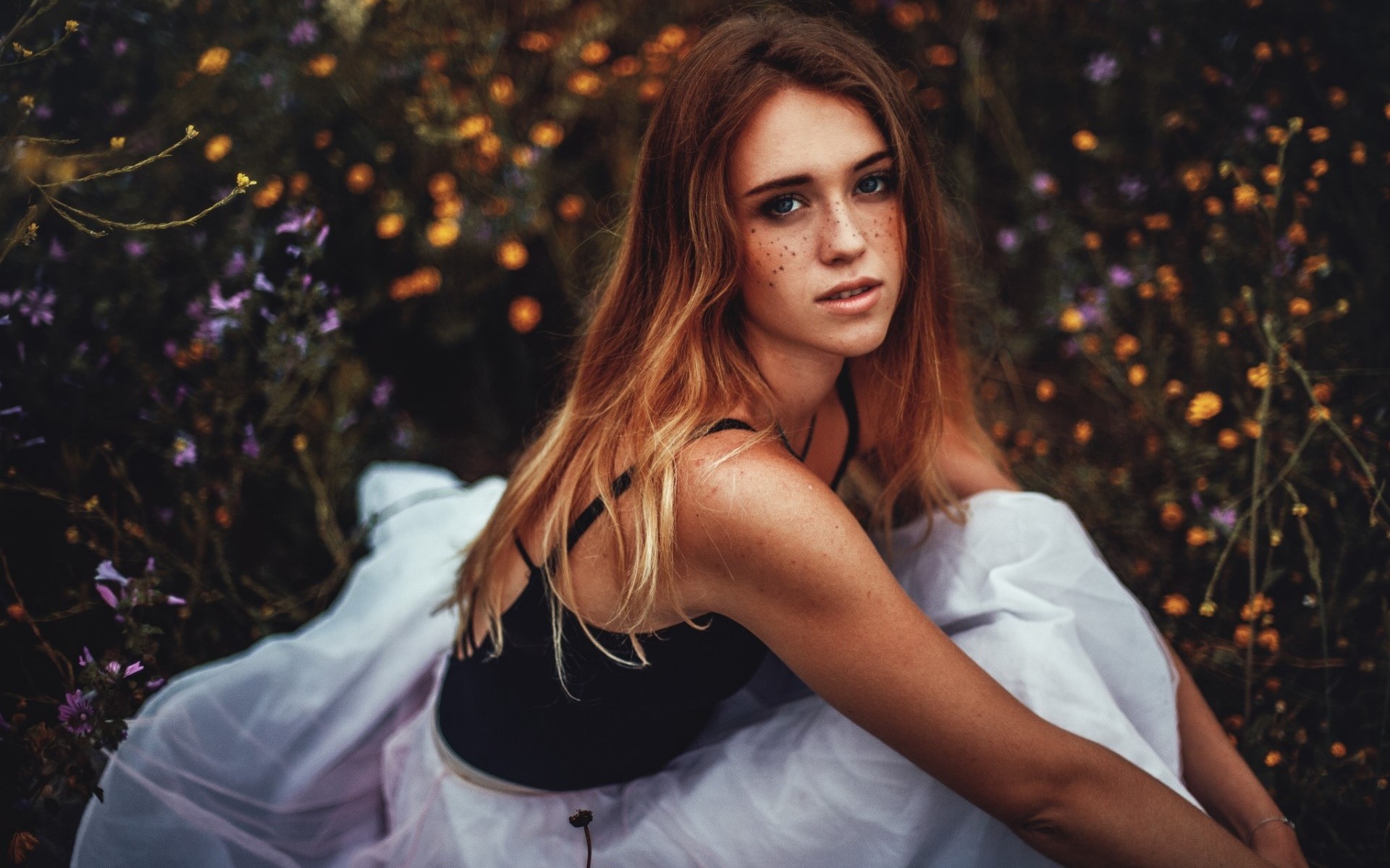 Woman with freckles sitting in a field of flowers HD Wallpaper