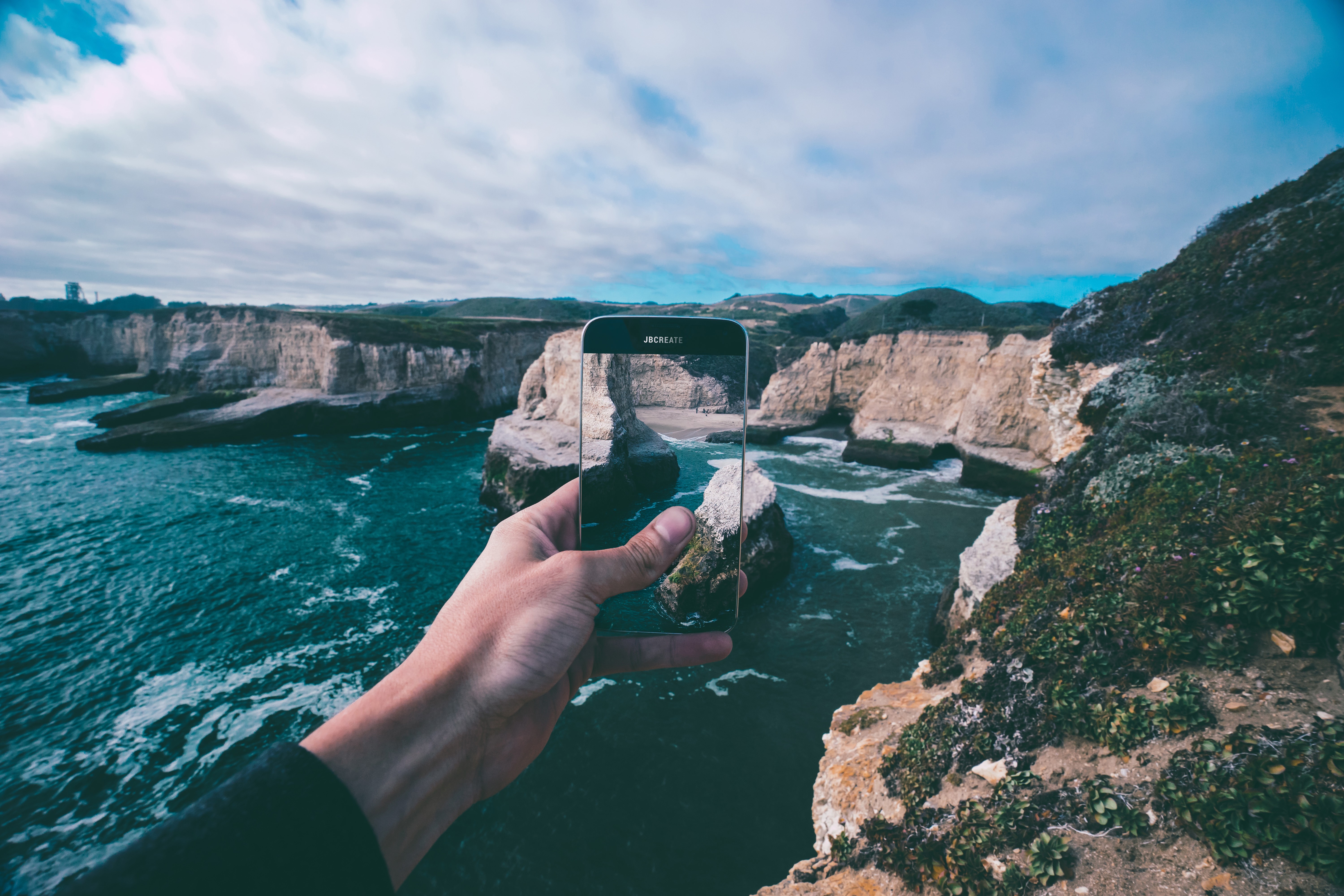 Hand holding phone capturing ocean cliffs and waves HD Wallpaper