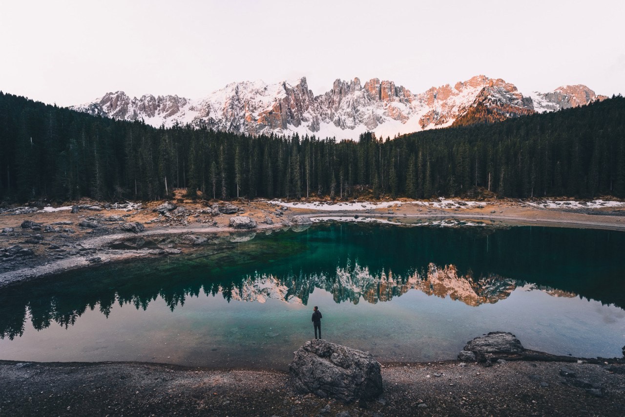 Man standing by a lake with snowy mountains and forest reflection HD Wallpaper