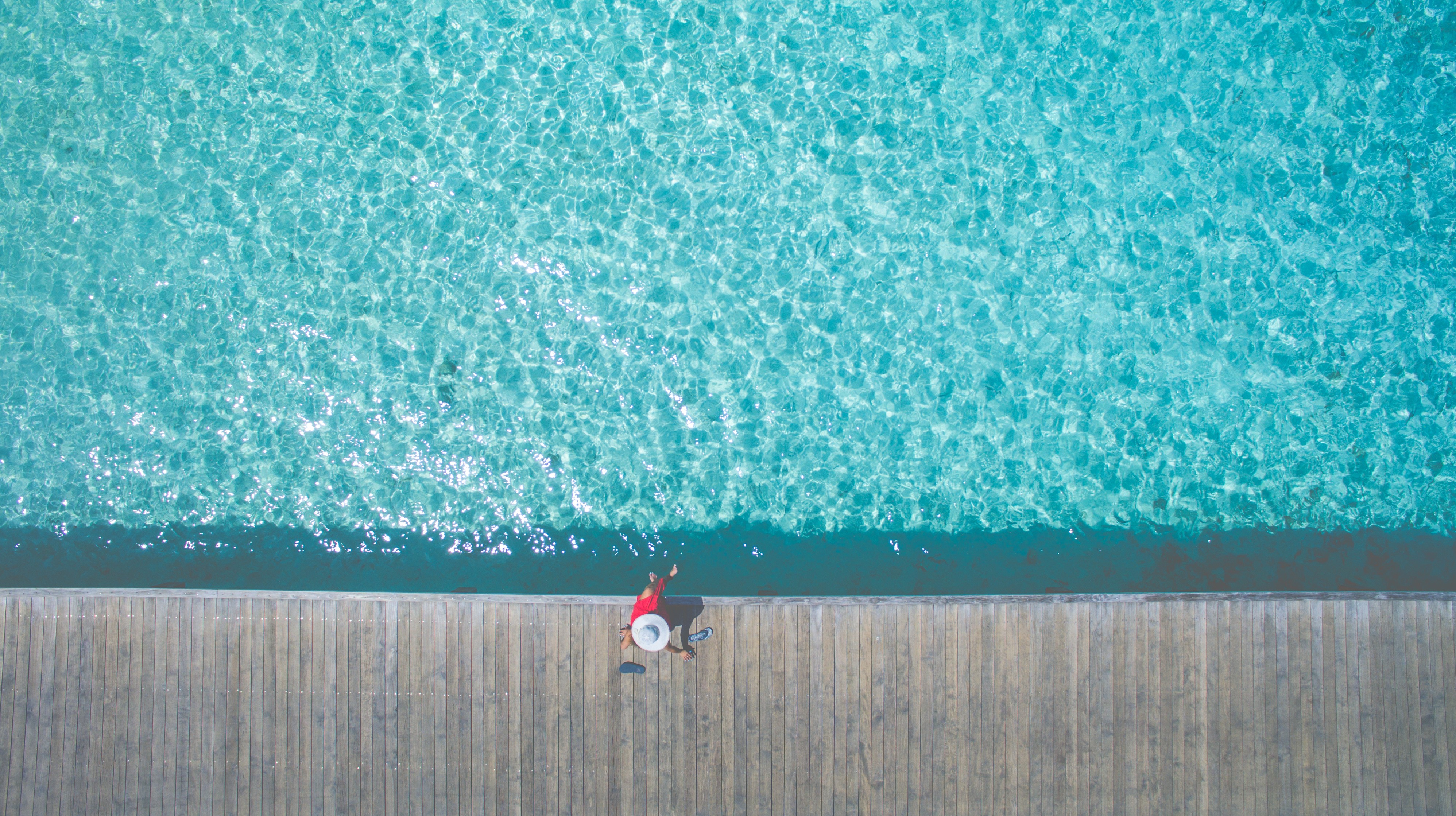 Person sitting on a wooden deck by clear blue water HD Wallpaper