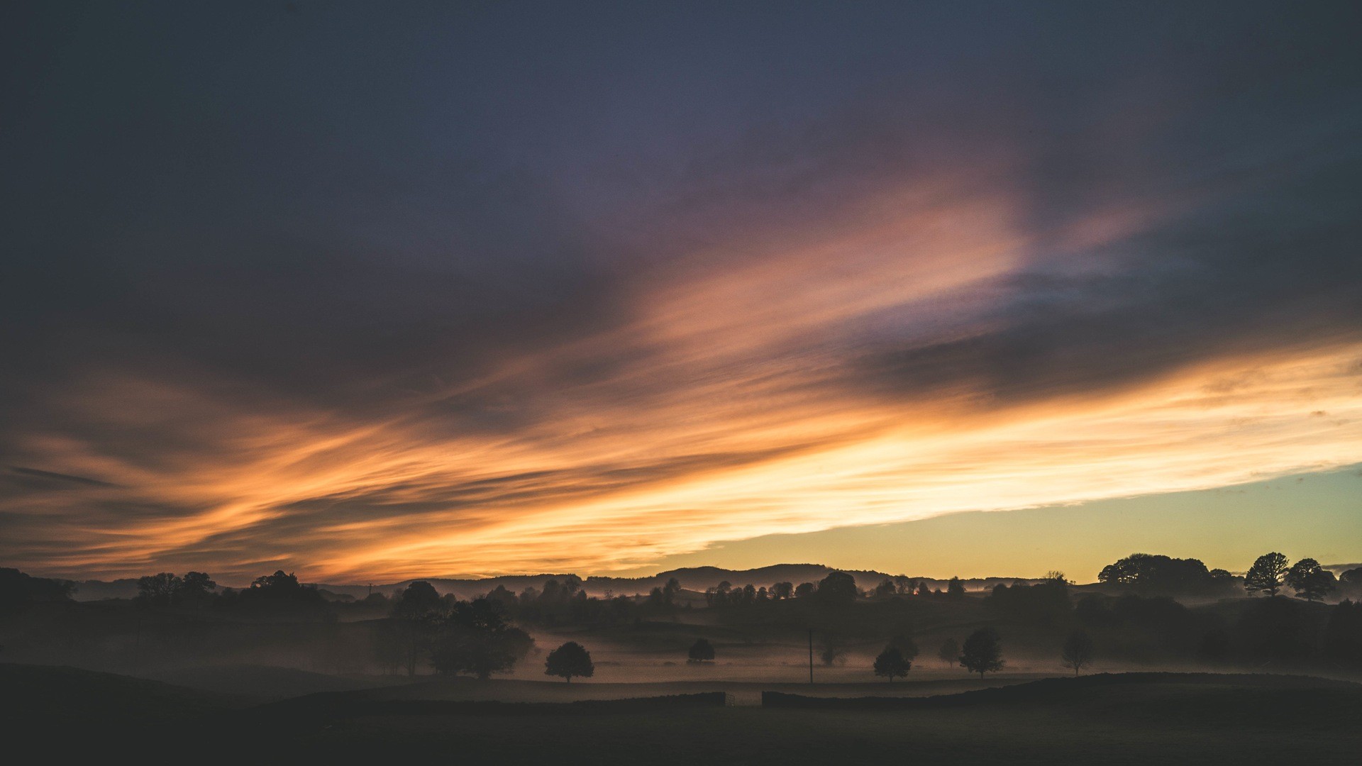 Misty sunrise over rolling hills with dramatic clouds HD Wallpaper