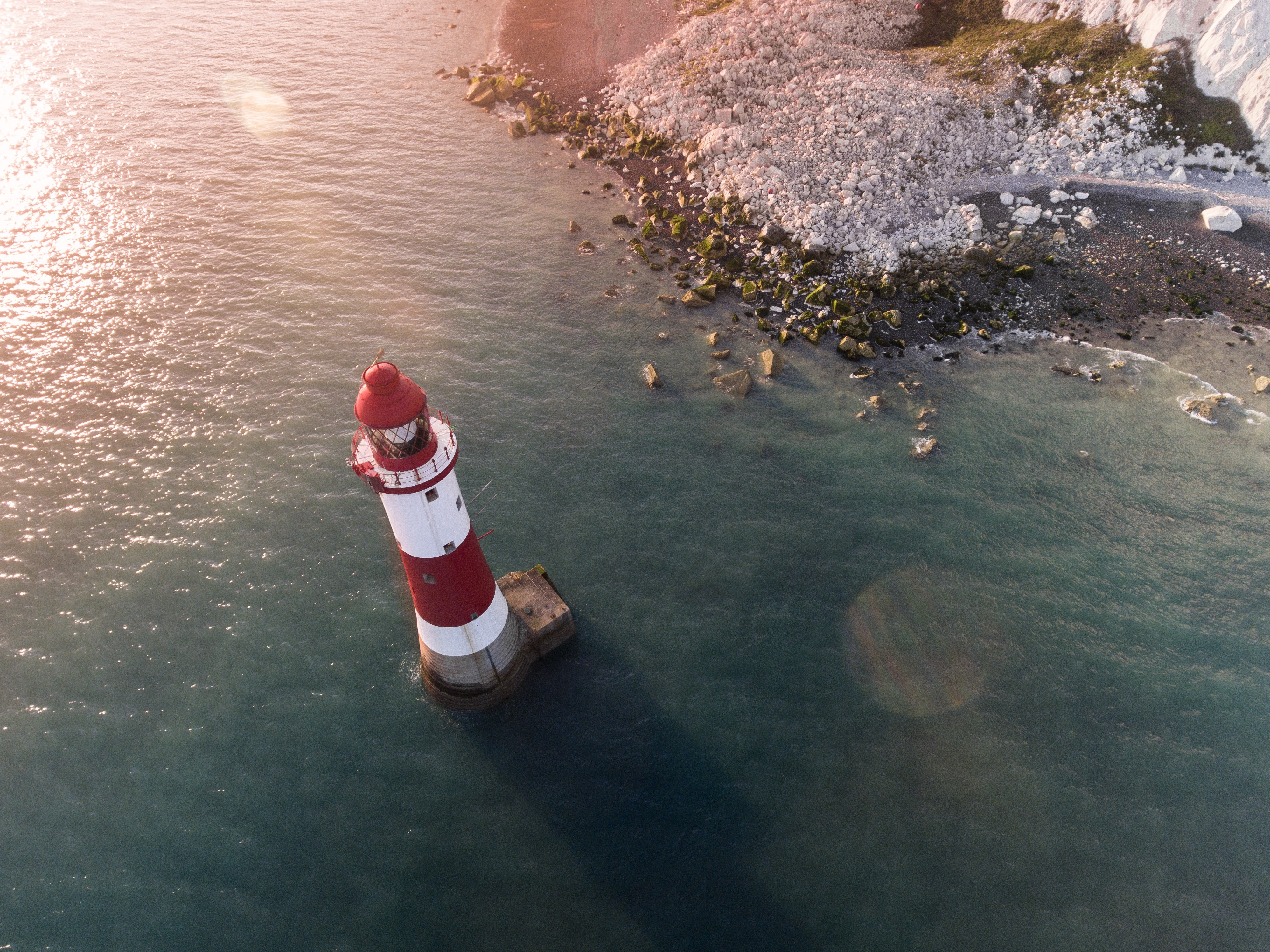 Red and white lighthouse on the water near a rocky shore HD Wallpaper