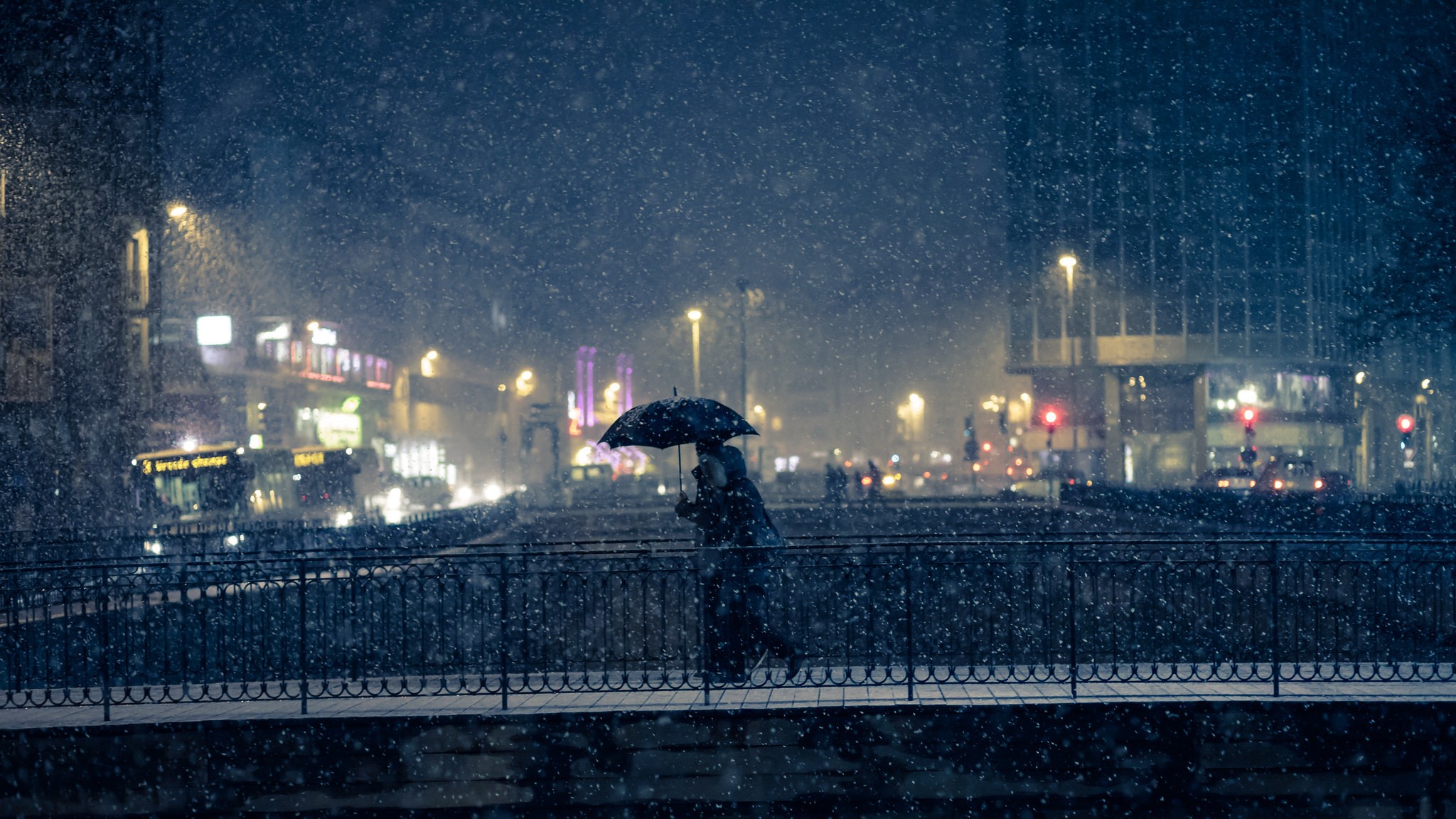 Person with umbrella walking on a bridge in a snowy city at night HD Wallpaper