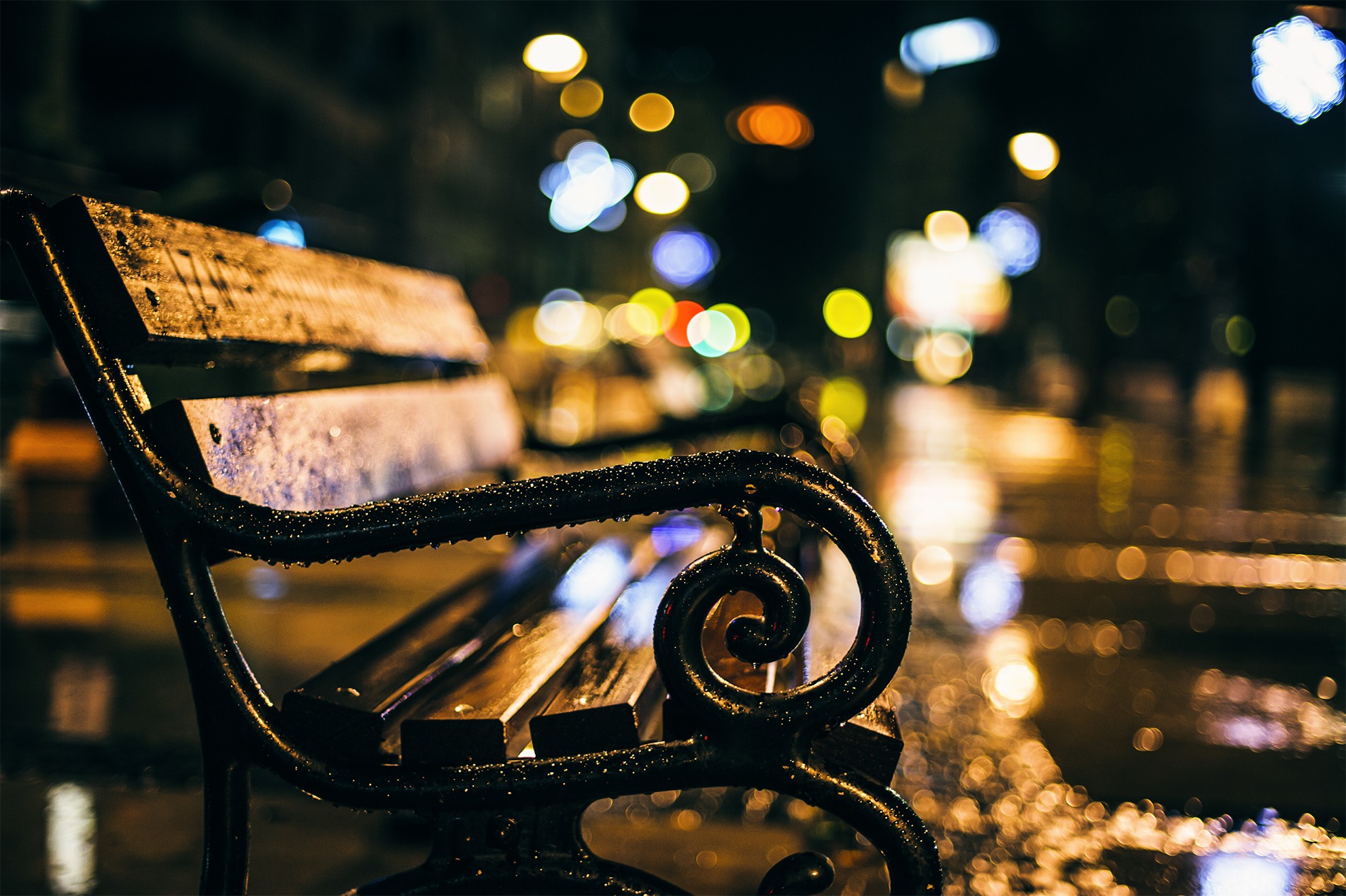 A wet park bench at night with city lights in the background HD Wallpaper