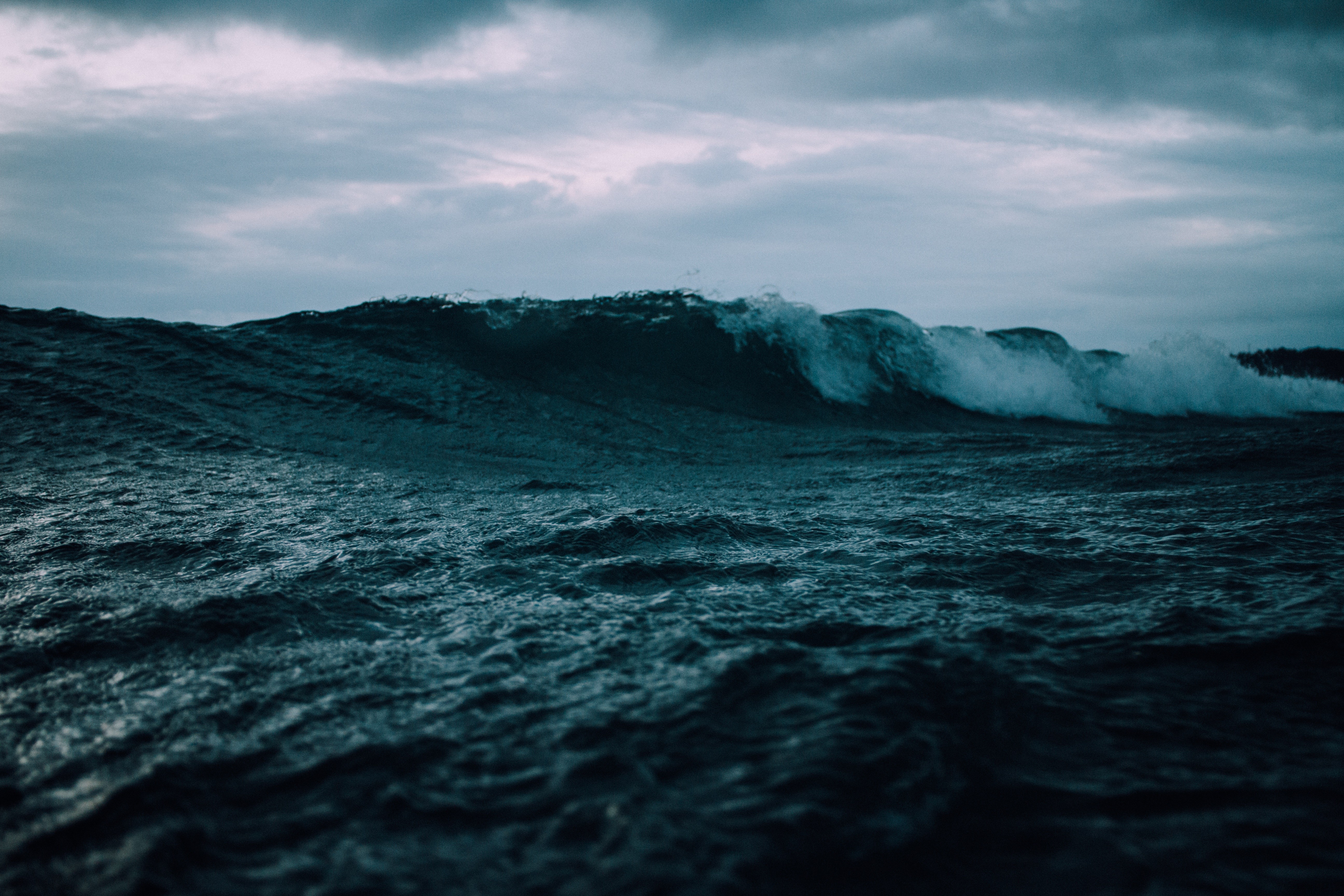 A large wave crashes in the dark ocean under a cloudy sky. HD Wallpaper