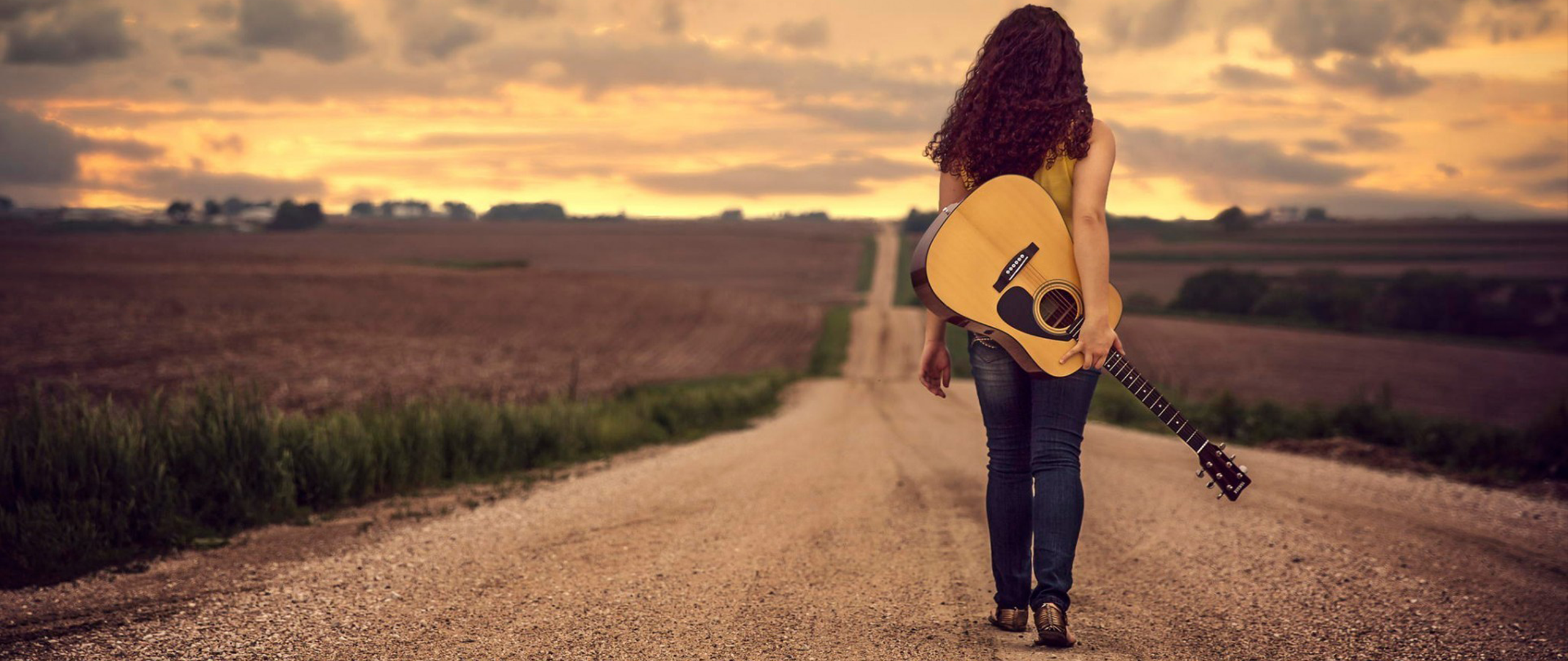Woman with guitar walking on a dirt road at sunset HD Wallpaper