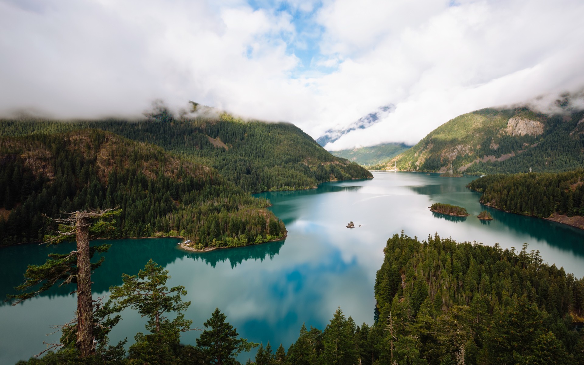 Turquoise lake surrounded by forested mountains under clouds HD Wallpaper