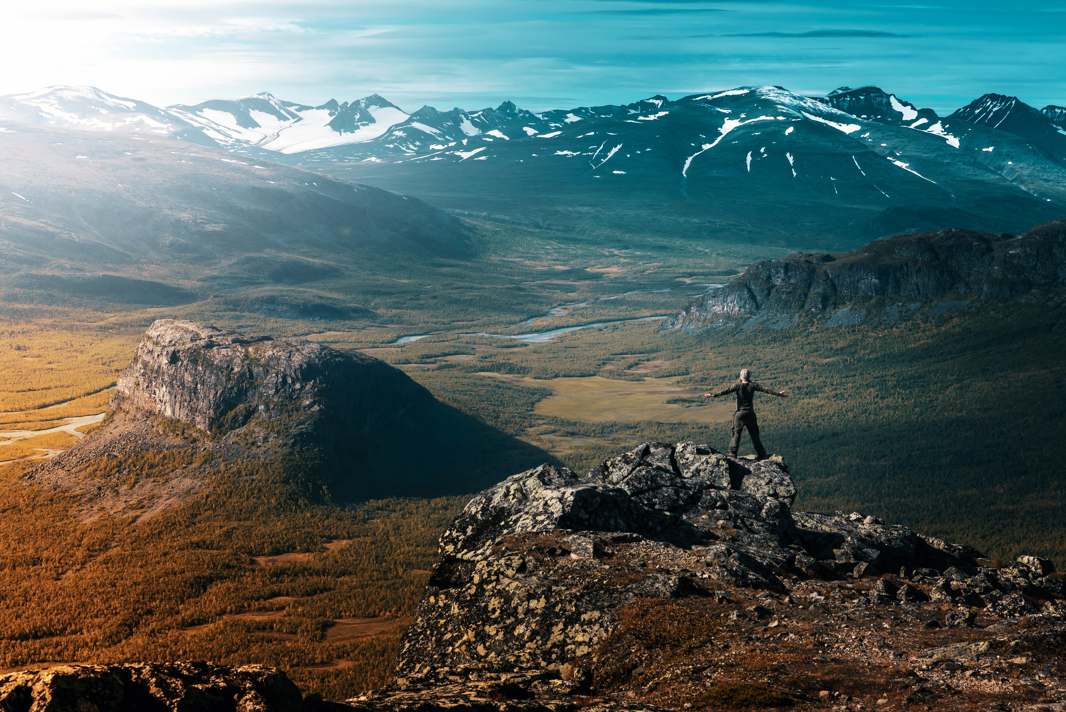 Person standing on a rocky outcrop overlooking a vast mountain valley HD Wallpaper