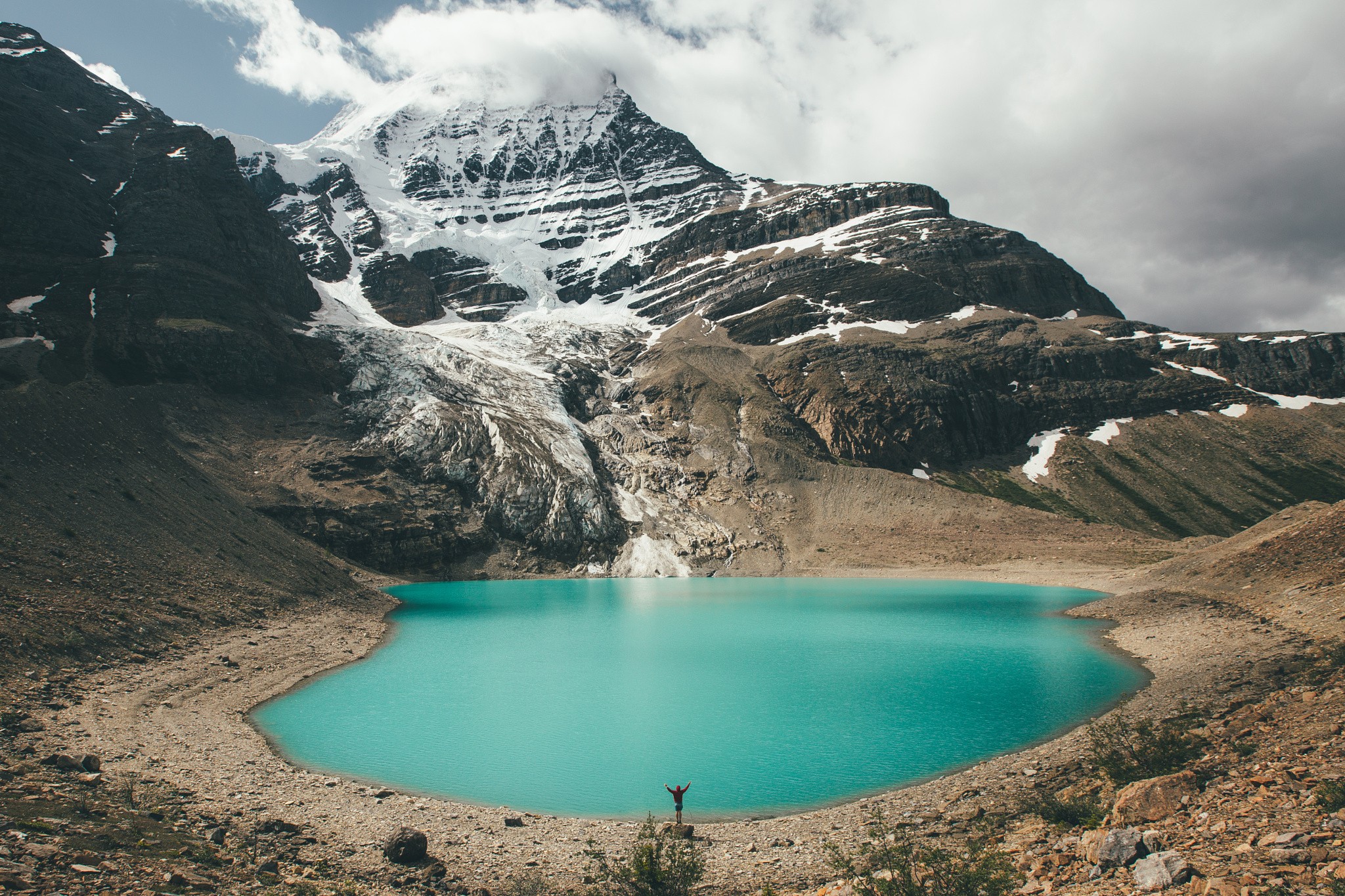 Person standing by a turquoise lake with snow-capped mountains HD Wallpaper