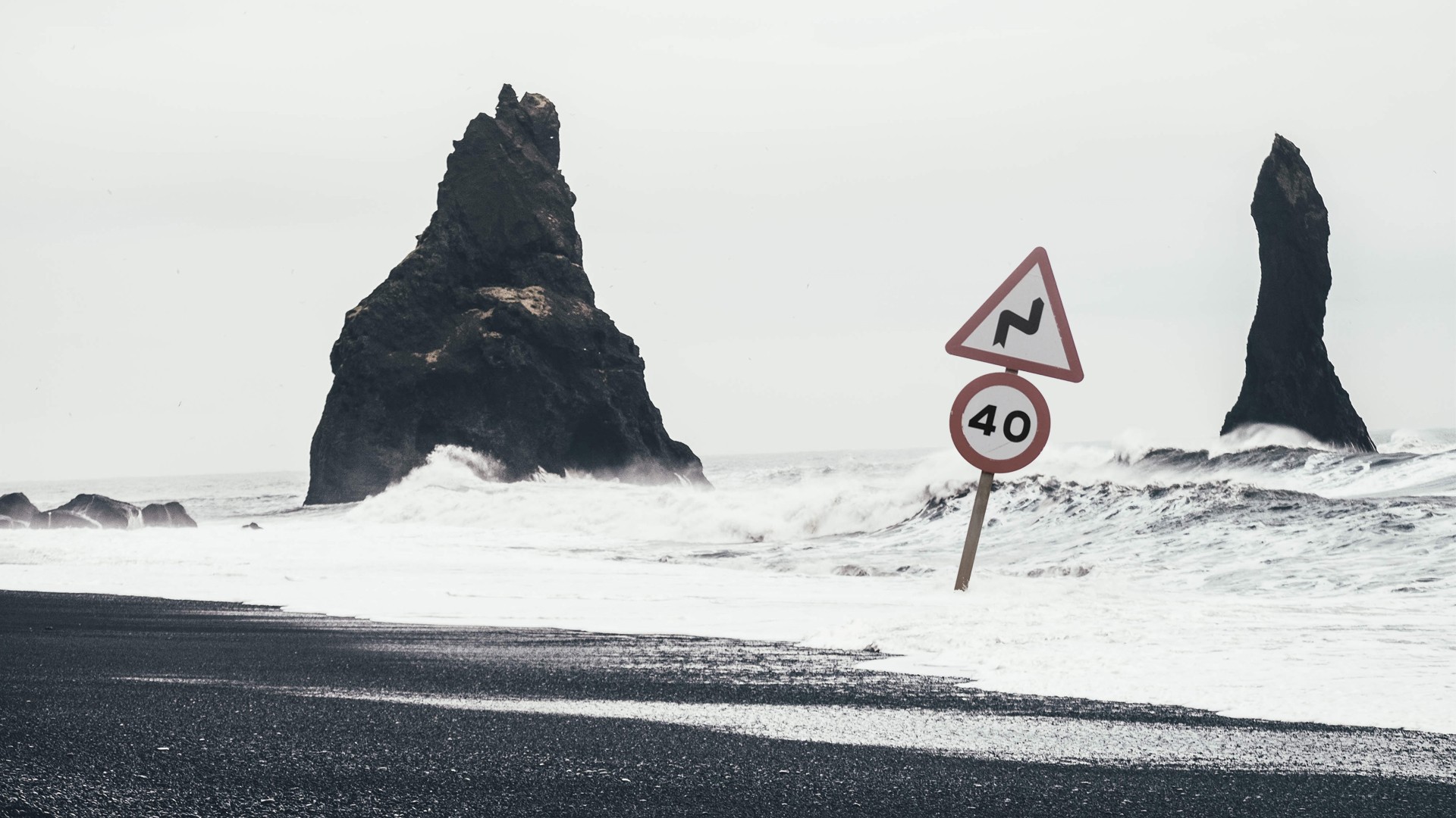 Road signs on a black sand beach with waves and rock formations HD Wallpaper