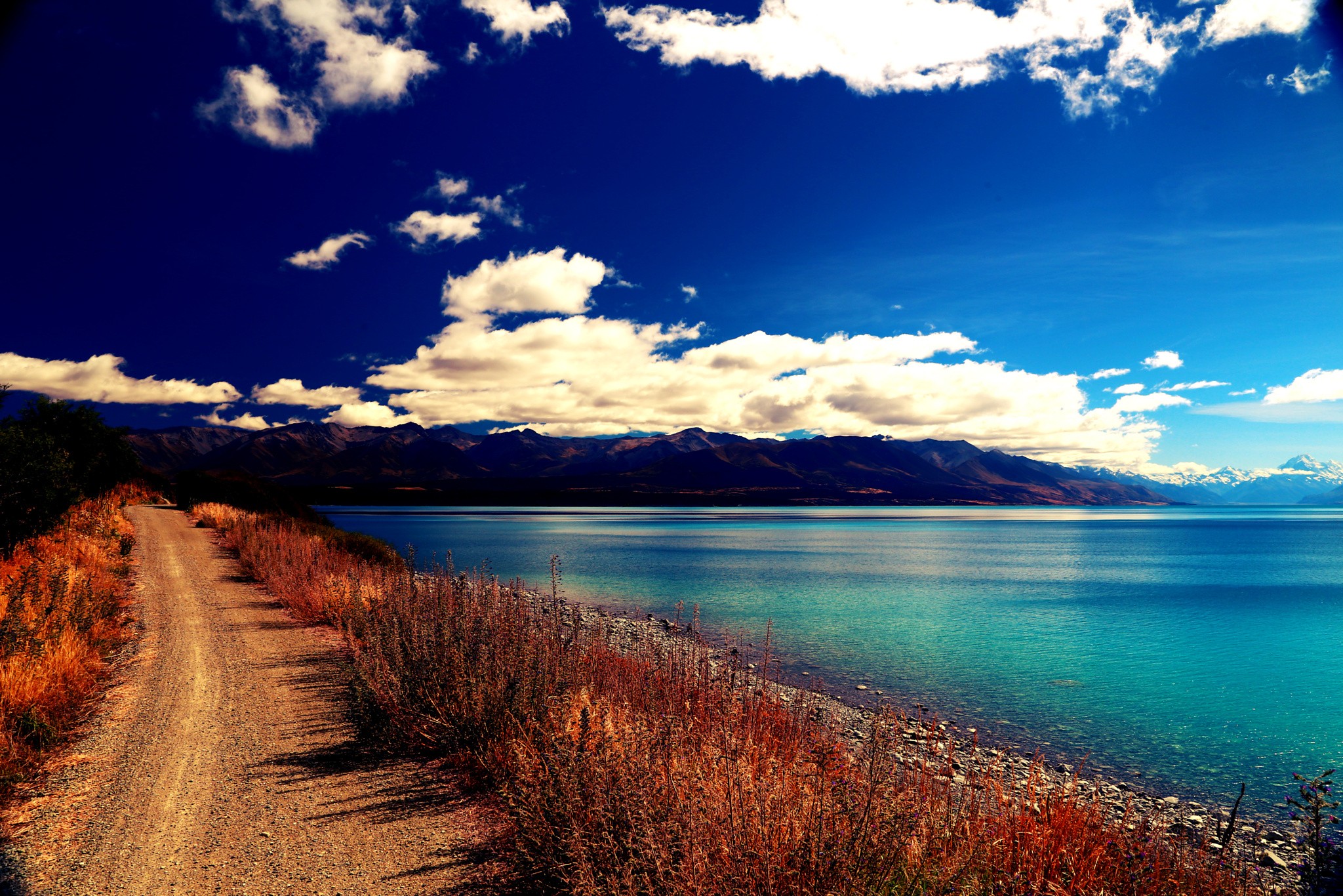 Dirt road beside a turquoise lake and mountains under a blue sky HD Wallpaper
