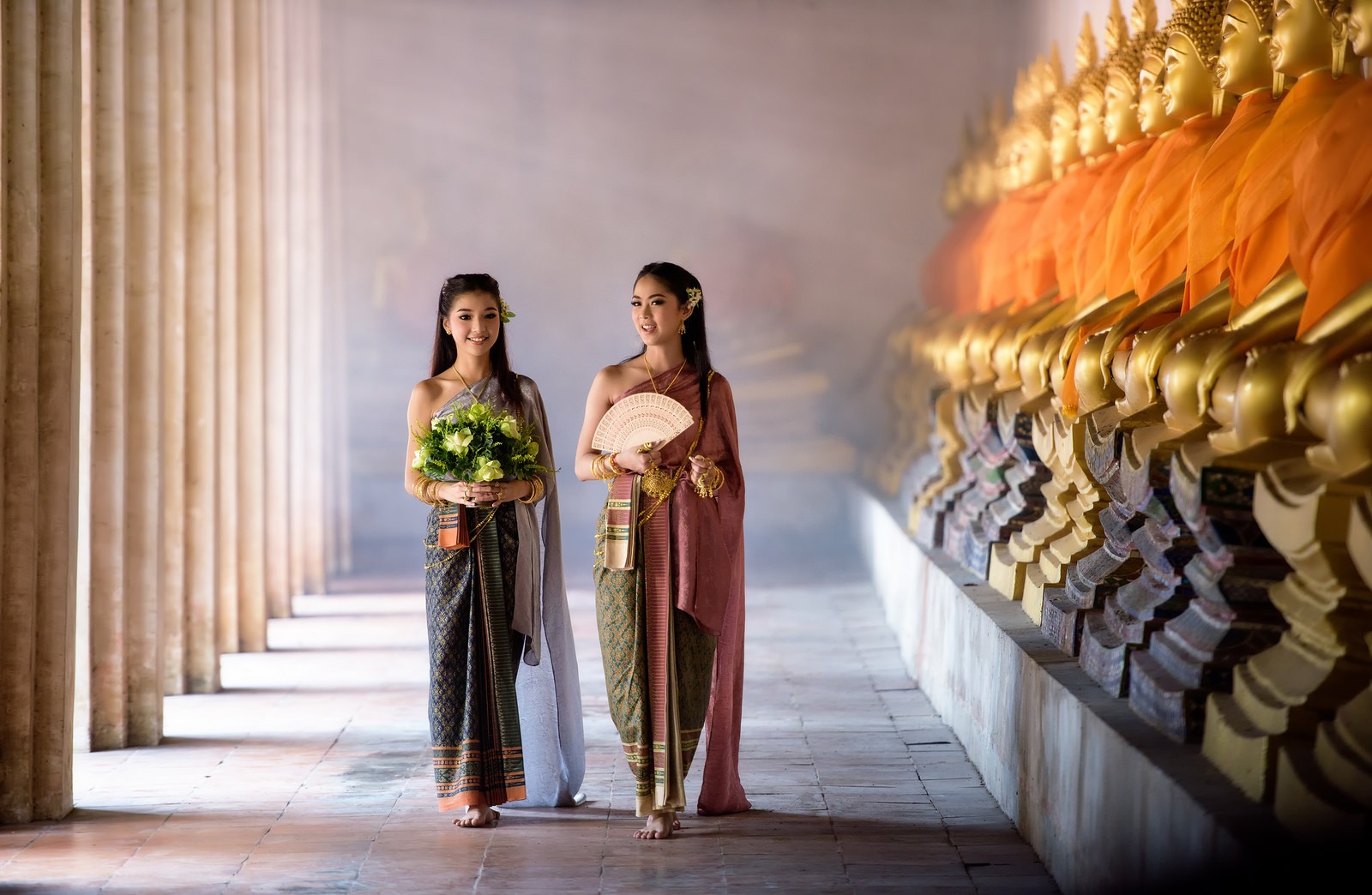 Two women in traditional Thai clothing walking near Buddha statues HD Wallpaper