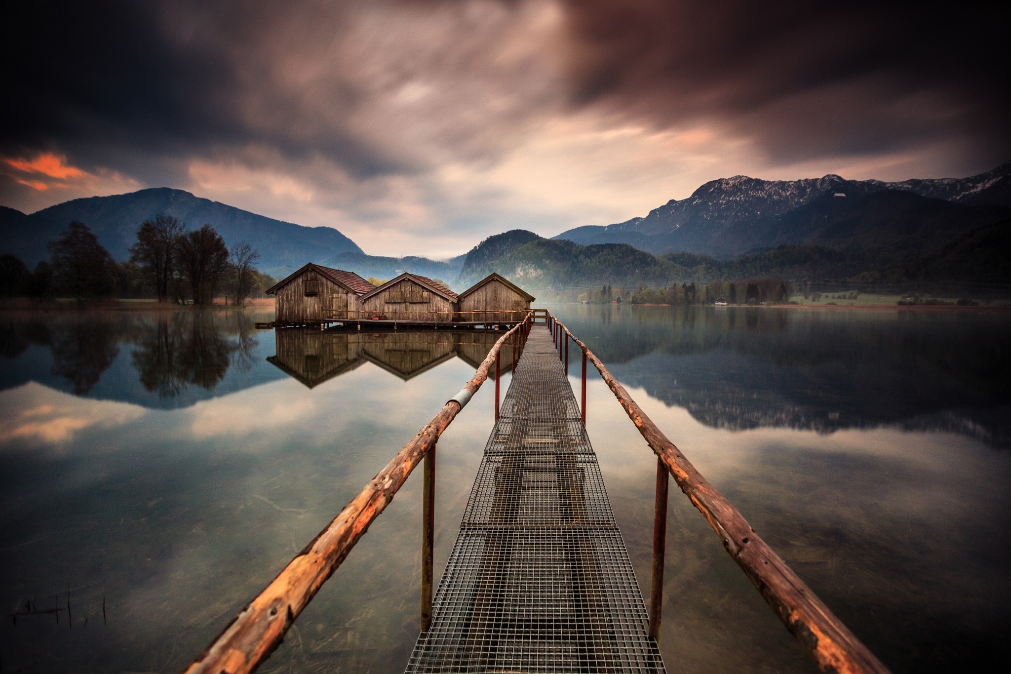 Wooden boathouses on a calm lake with mountains and a pier HD Wallpaper