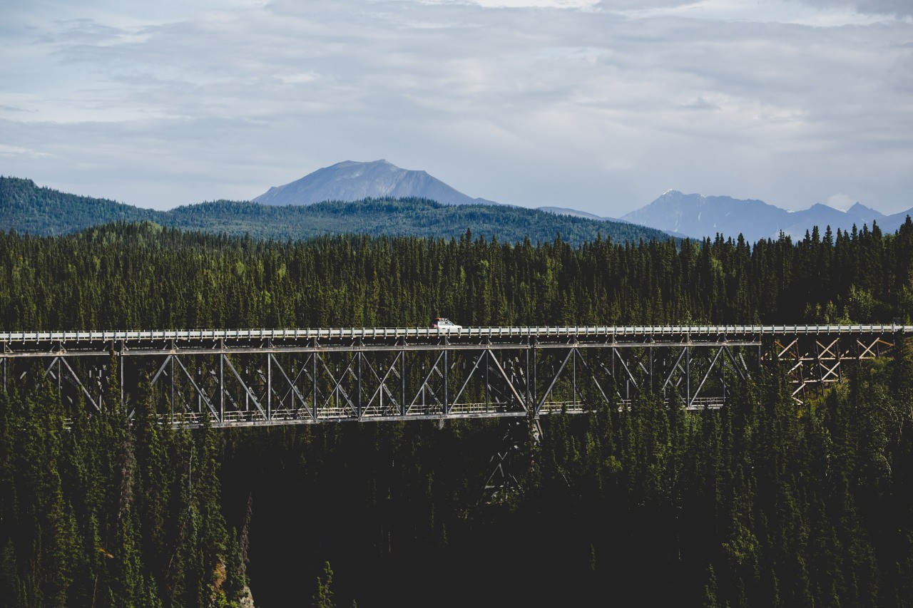 A white car drives across a bridge over a forest with mountains in the background. HD Wallpaper