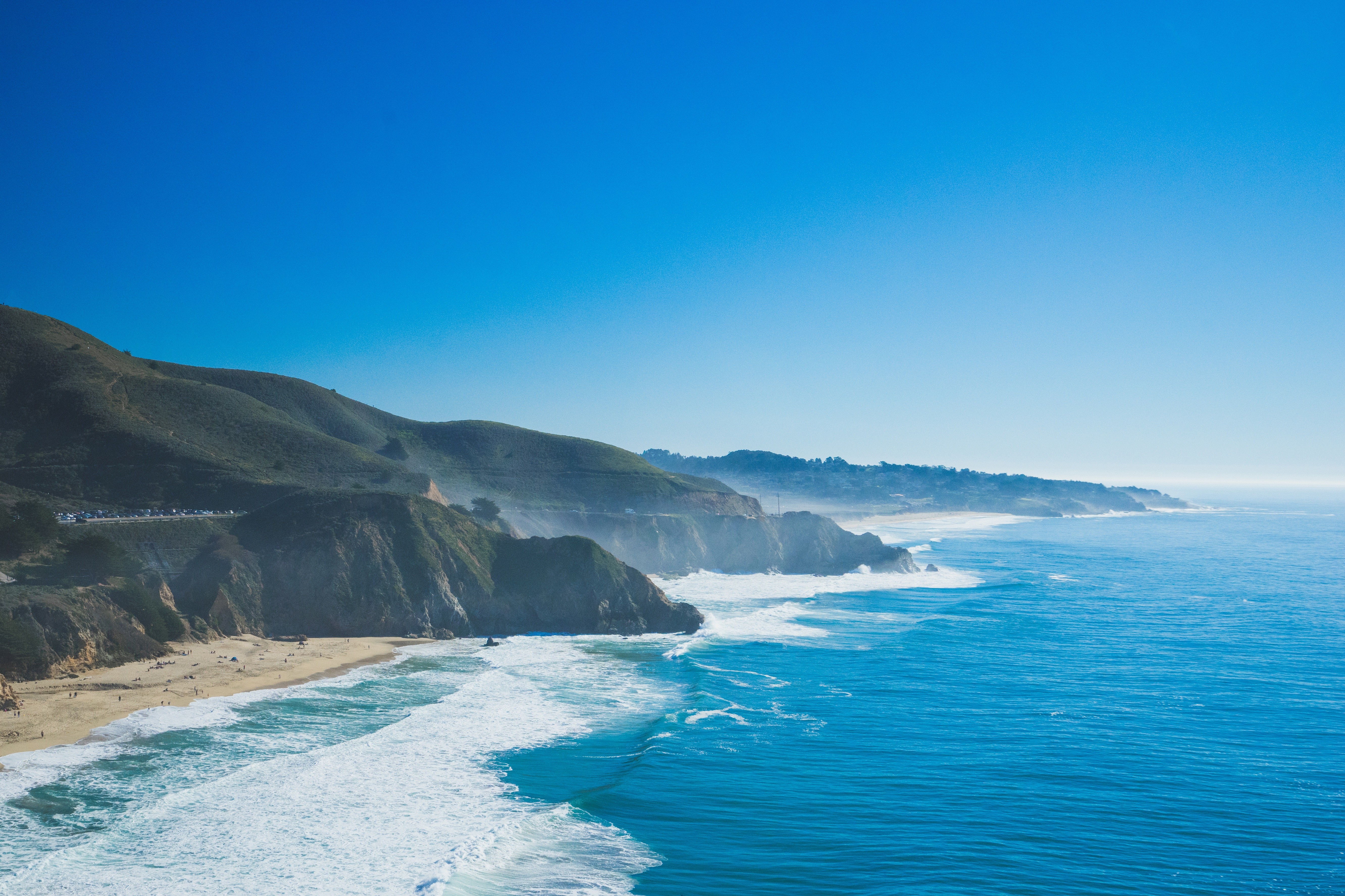 Ocean waves crash against a rugged coastline under a clear blue sky. HD Wallpaper