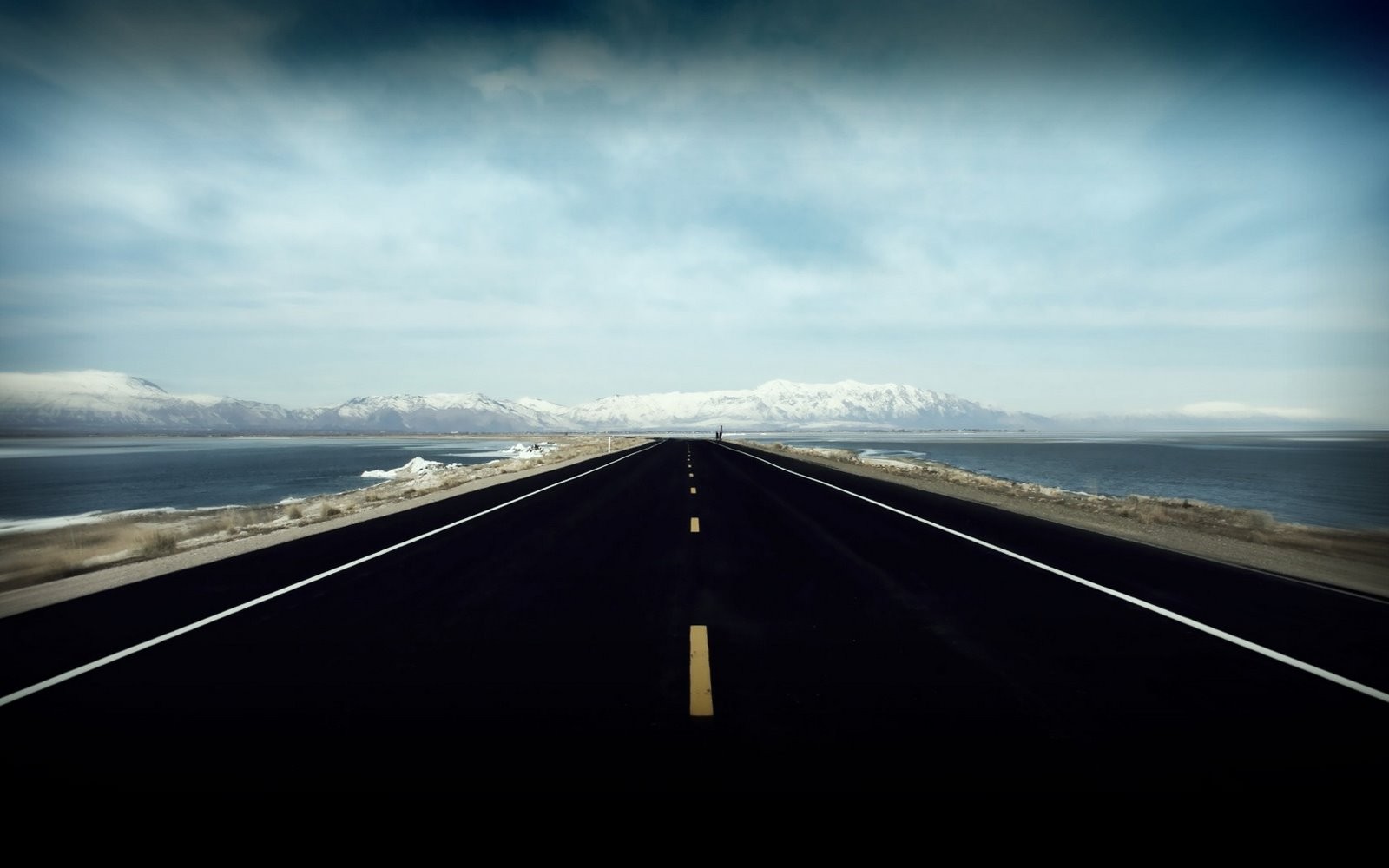 A long, empty road stretches towards snow-capped mountains. HD Wallpaper