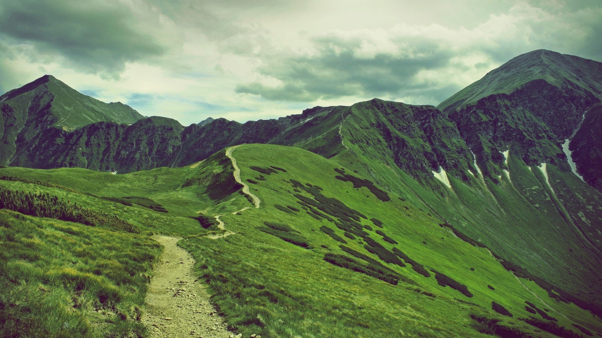 A winding path through green mountains under a cloudy sky HD Wallpaper