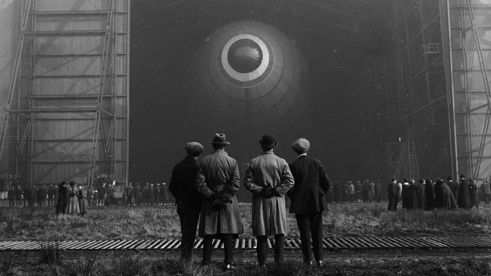 Men in coats and hats watch a Zeppelin in a hangar HD Wallpaper