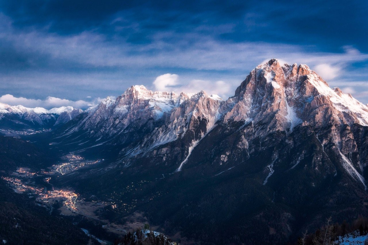 Snow-capped mountains overlooking a valley town at dusk HD Wallpaper