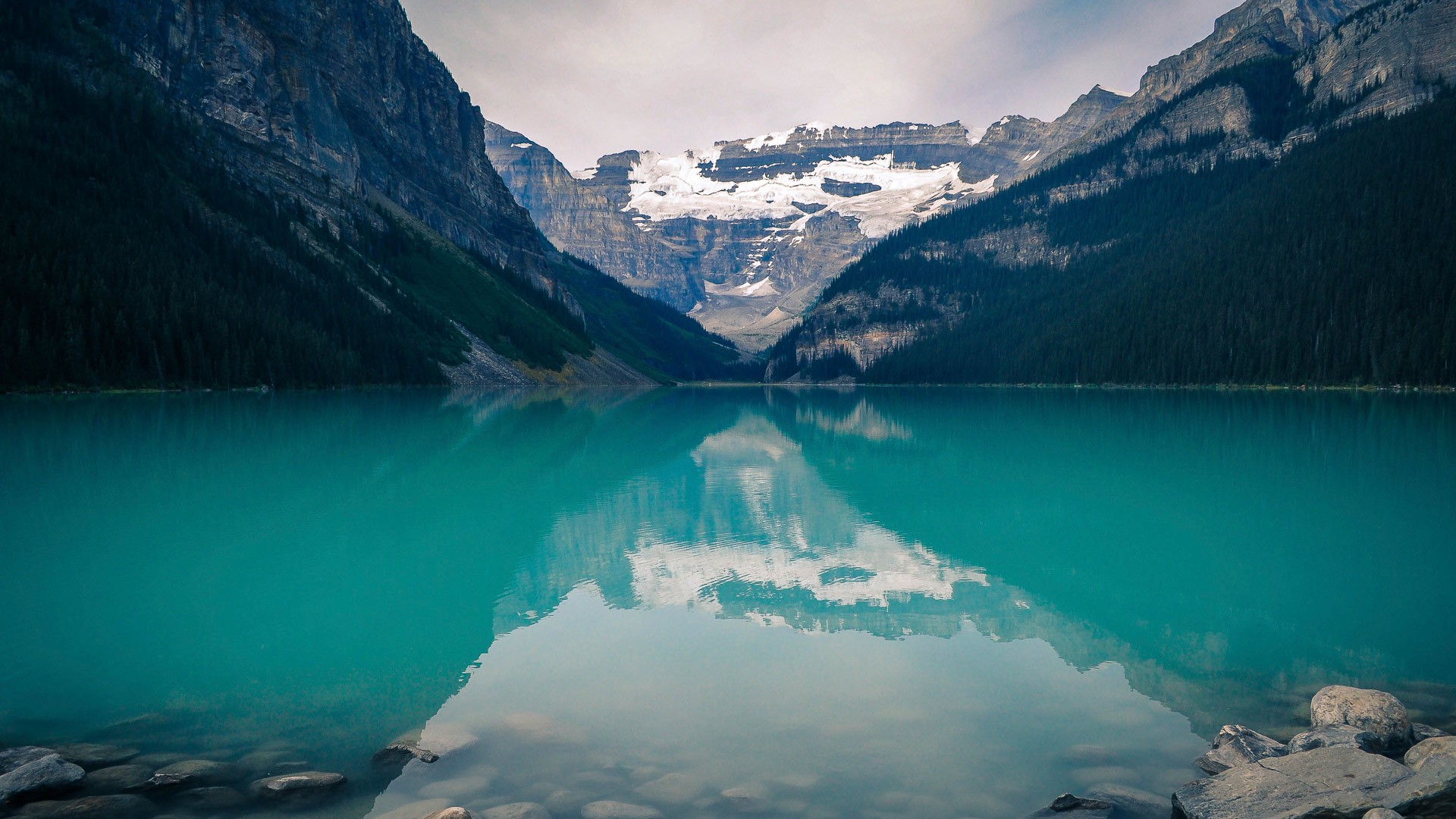 Turquoise lake reflecting snow-capped mountains and dark forests HD Wallpaper