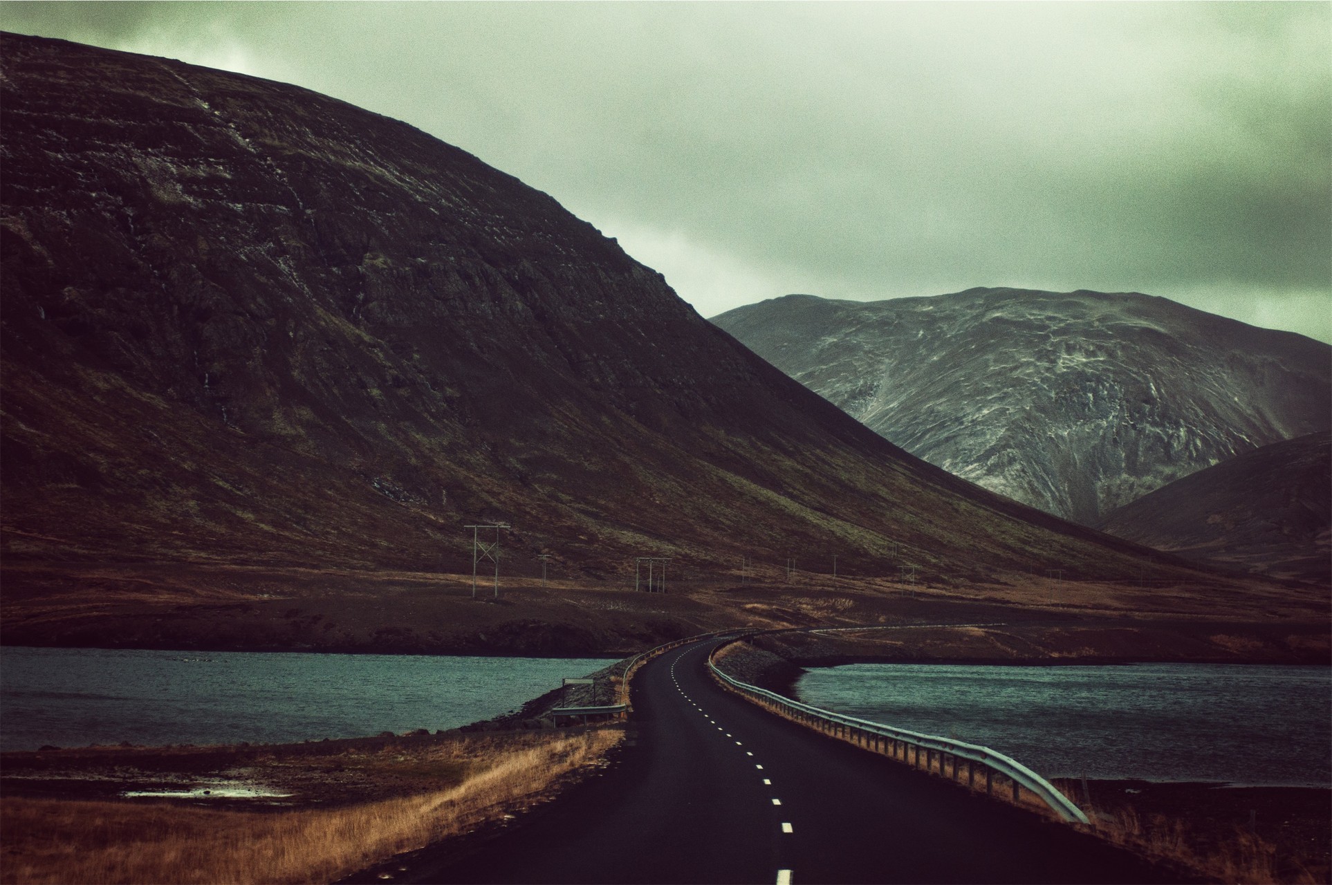 Road through mountains beside a lake under stormy clouds HD Wallpaper