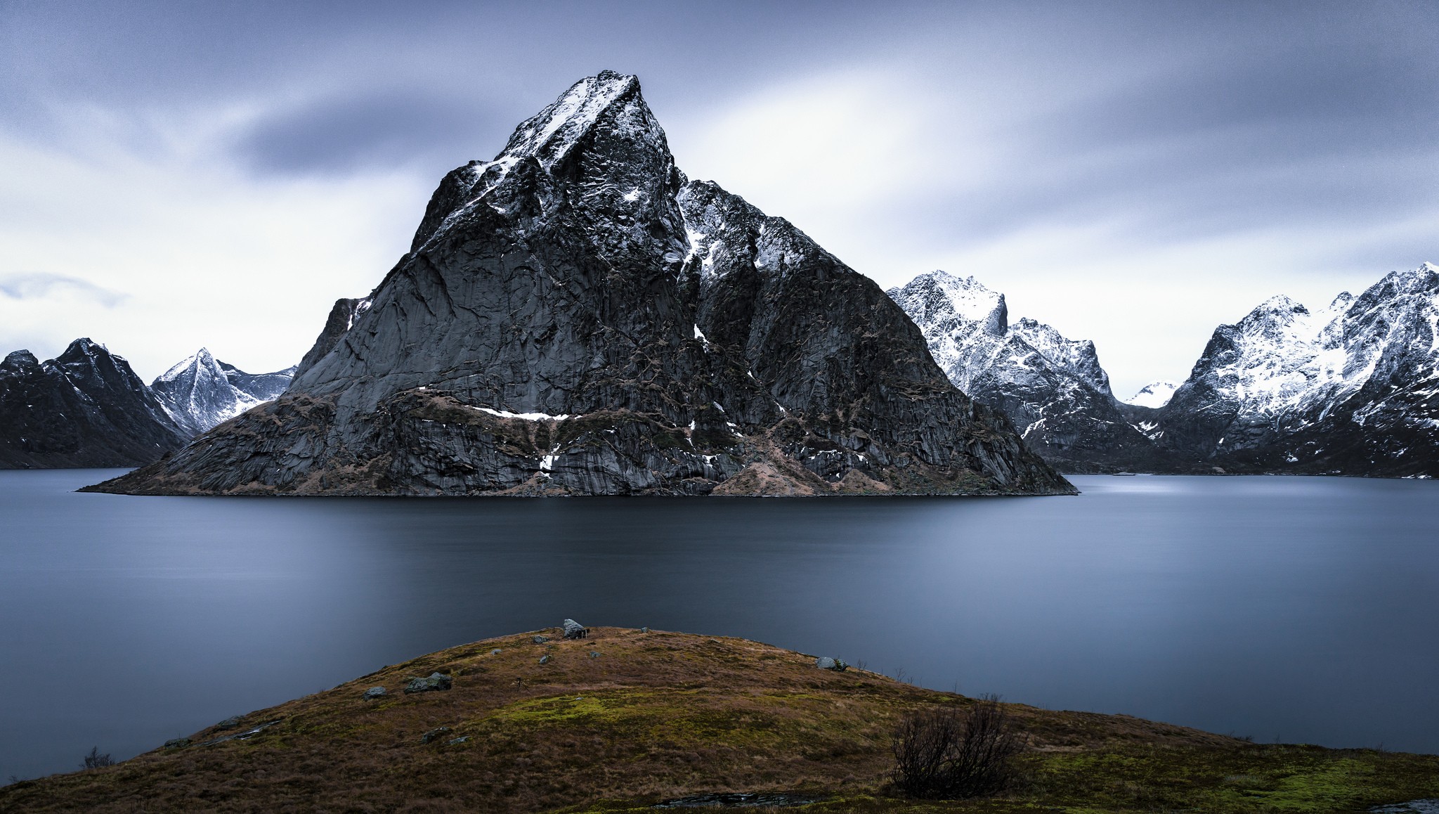 Snow-capped mountains rise from a calm lake under a cloudy sky HD Wallpaper