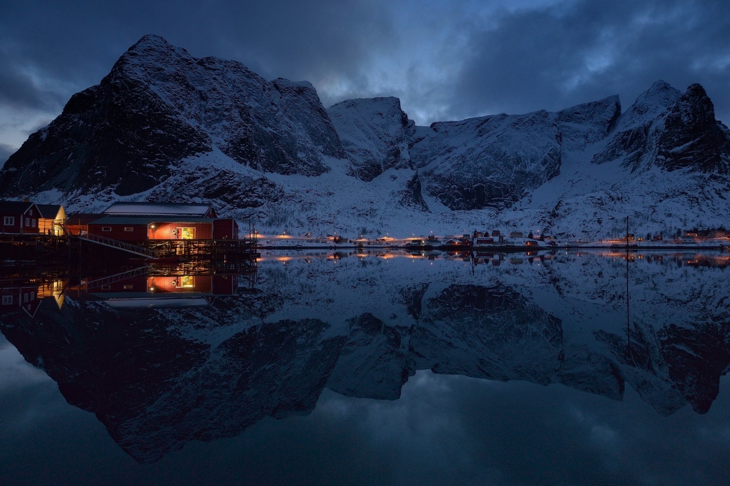 Snowy mountains reflected in a calm lake with village lights HD Wallpaper