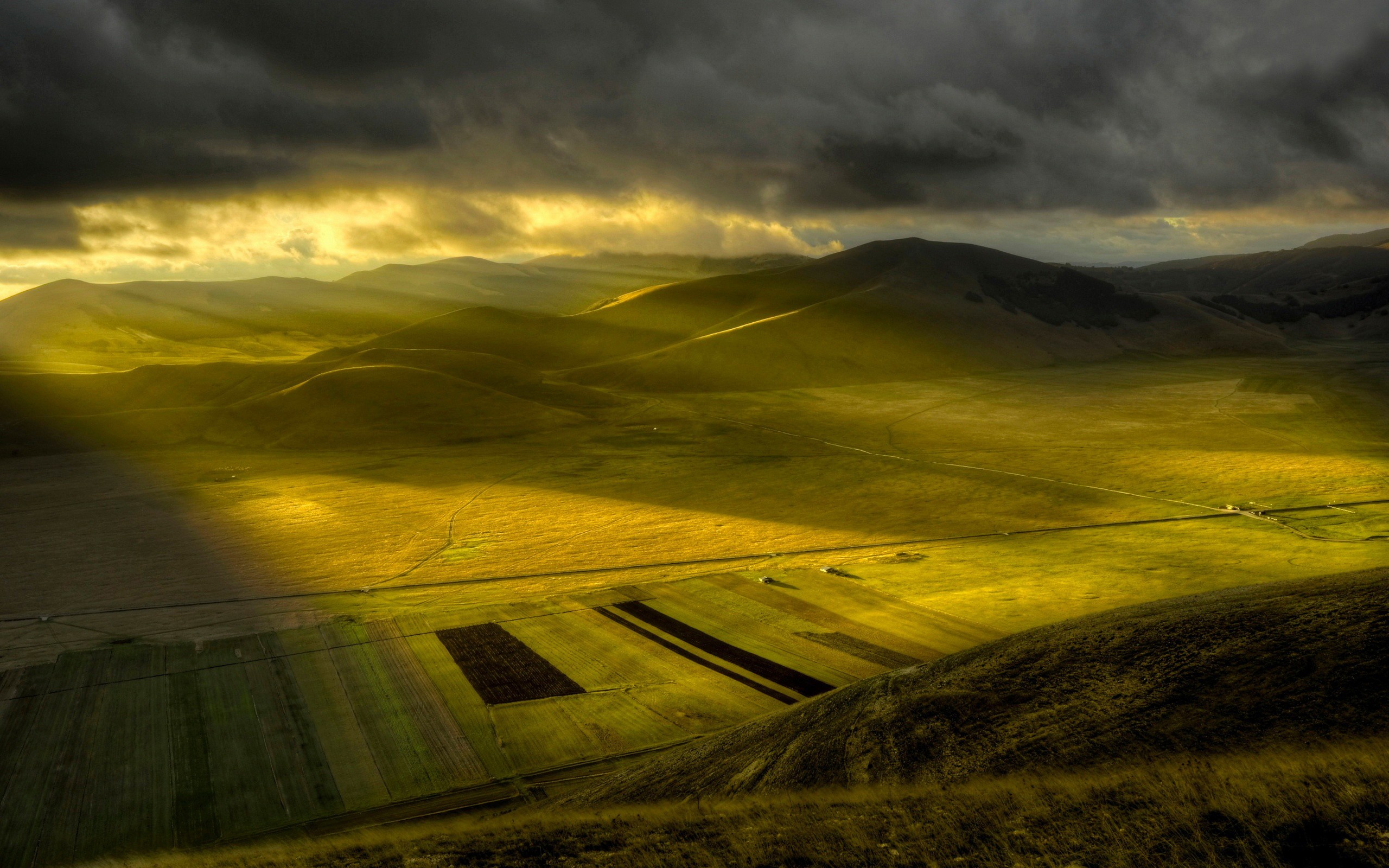 Sunlight beams through dark clouds over rolling hills and fields HD Wallpaper