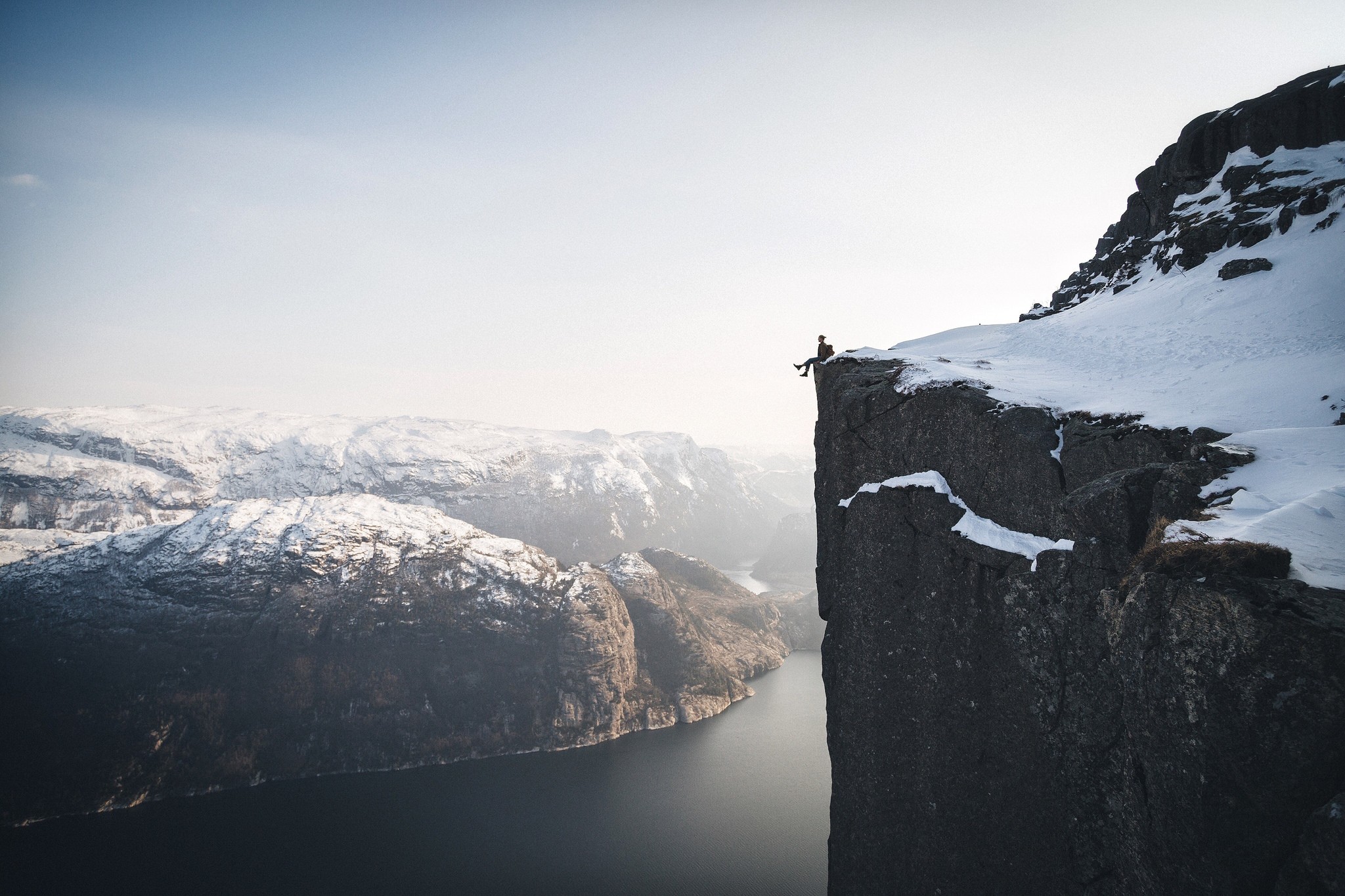 Person sitting on a snowy cliff overlooking a fjord in Norway HD Wallpaper