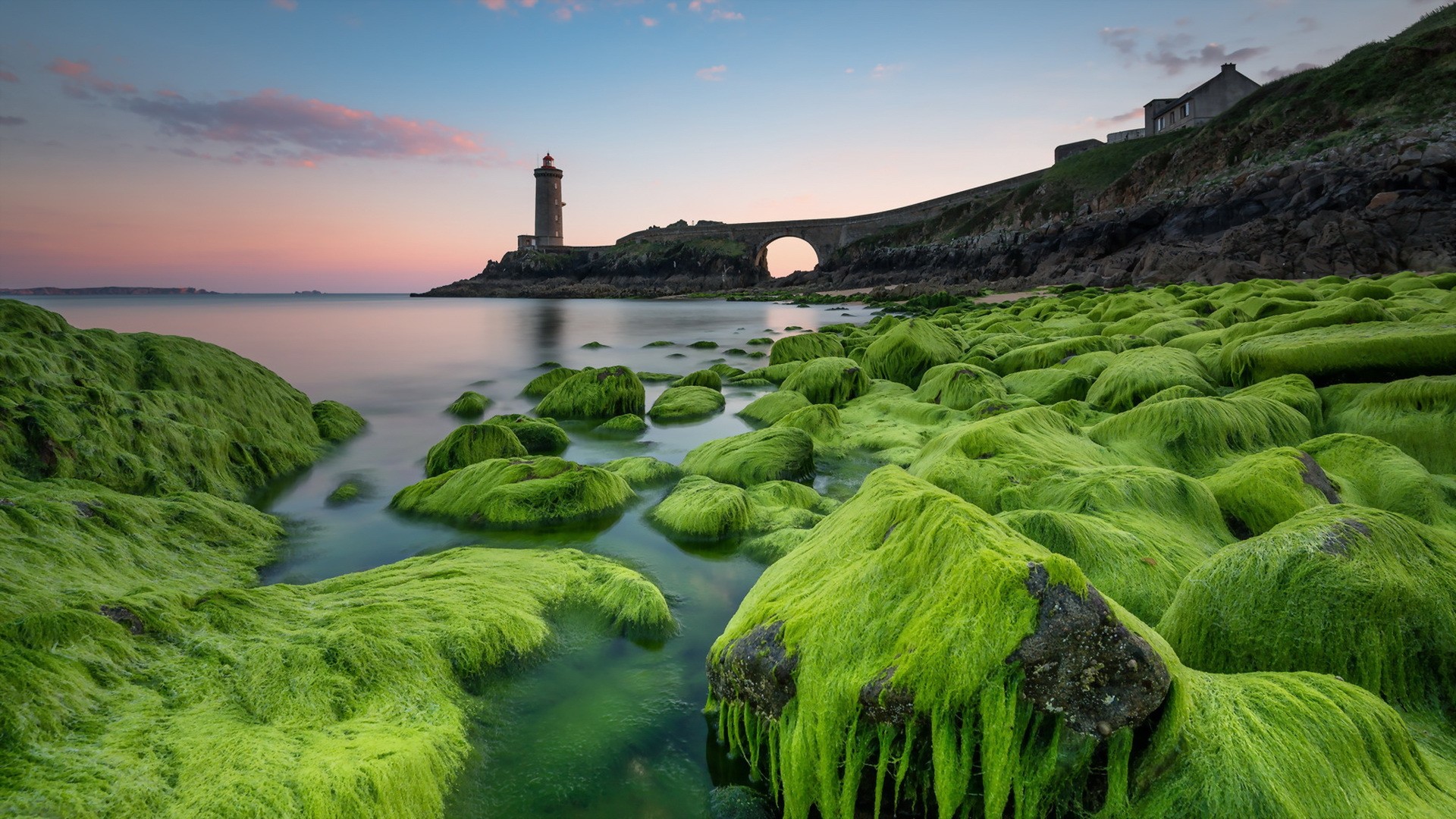 Lighthouse and bridge over moss-covered rocks at sunset HD Wallpaper