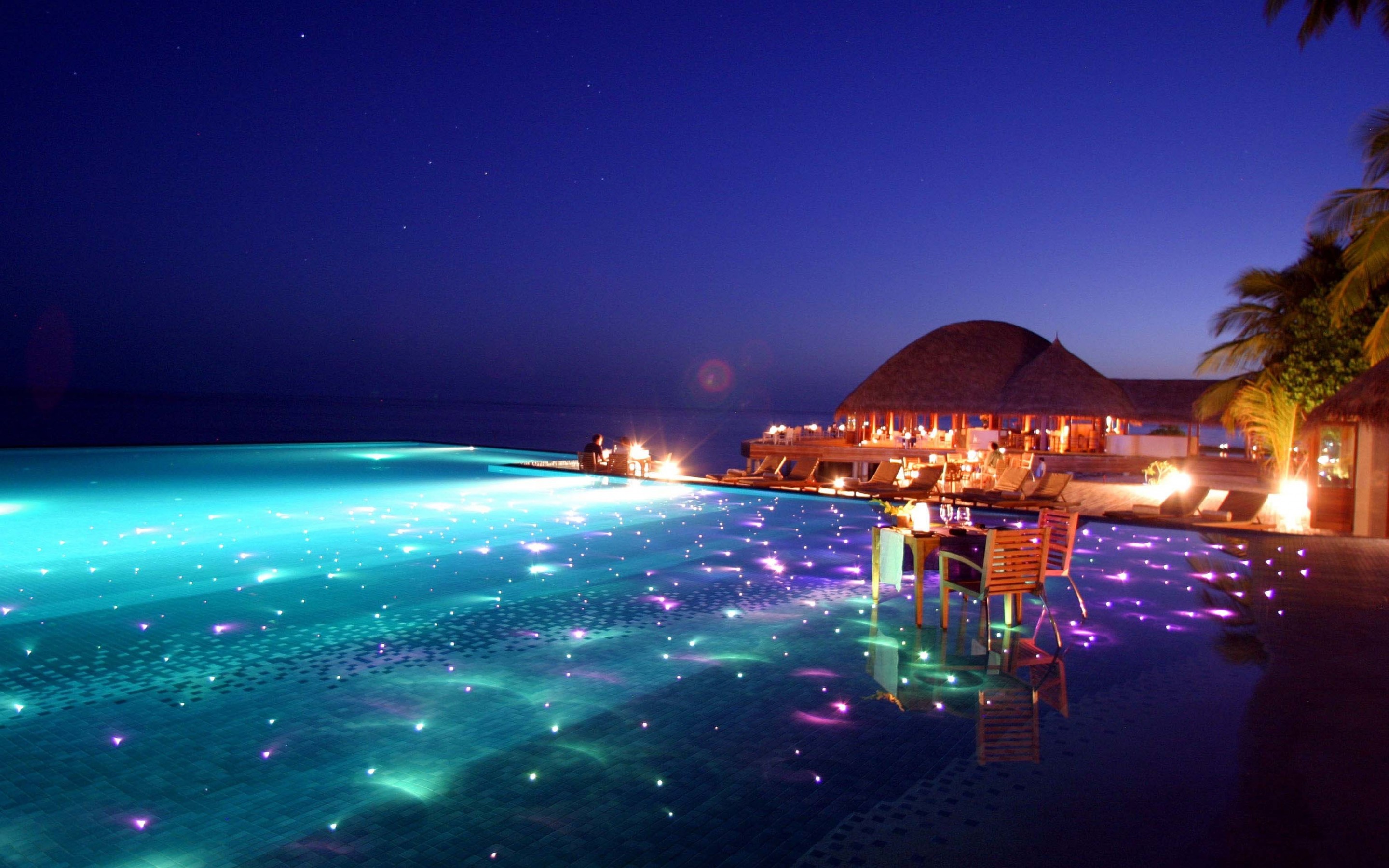 Infinity pool at night with glowing lights and a resort HD Wallpaper