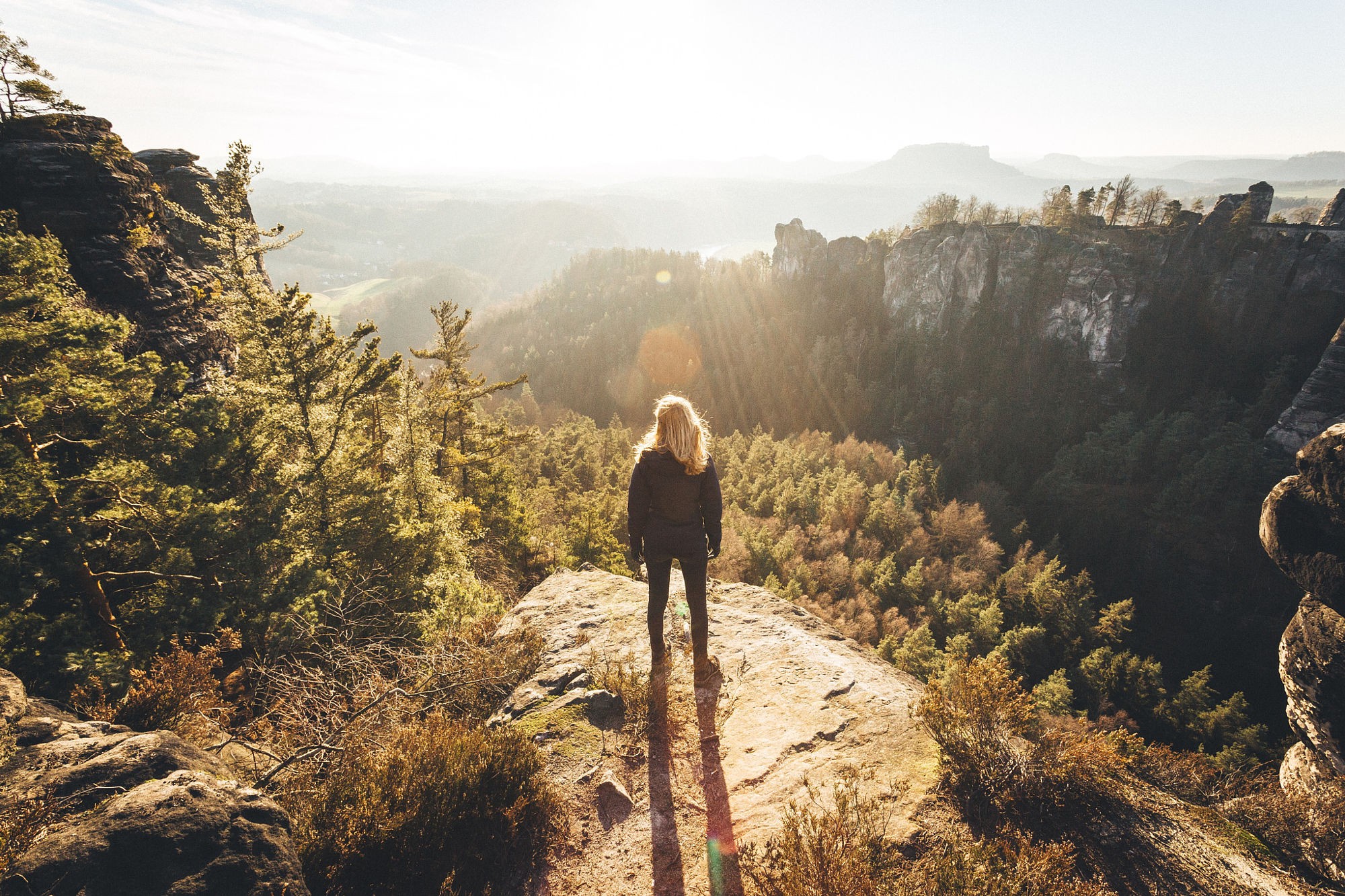 Woman standing on a cliff overlooking a forest landscape HD Wallpaper