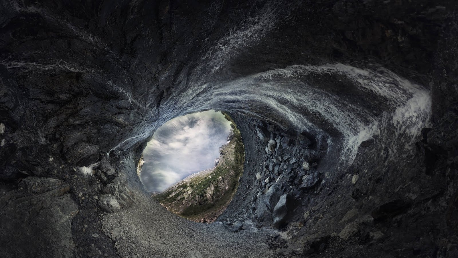 View from inside a dark cave looking out at mountains and clouds HD Wallpaper