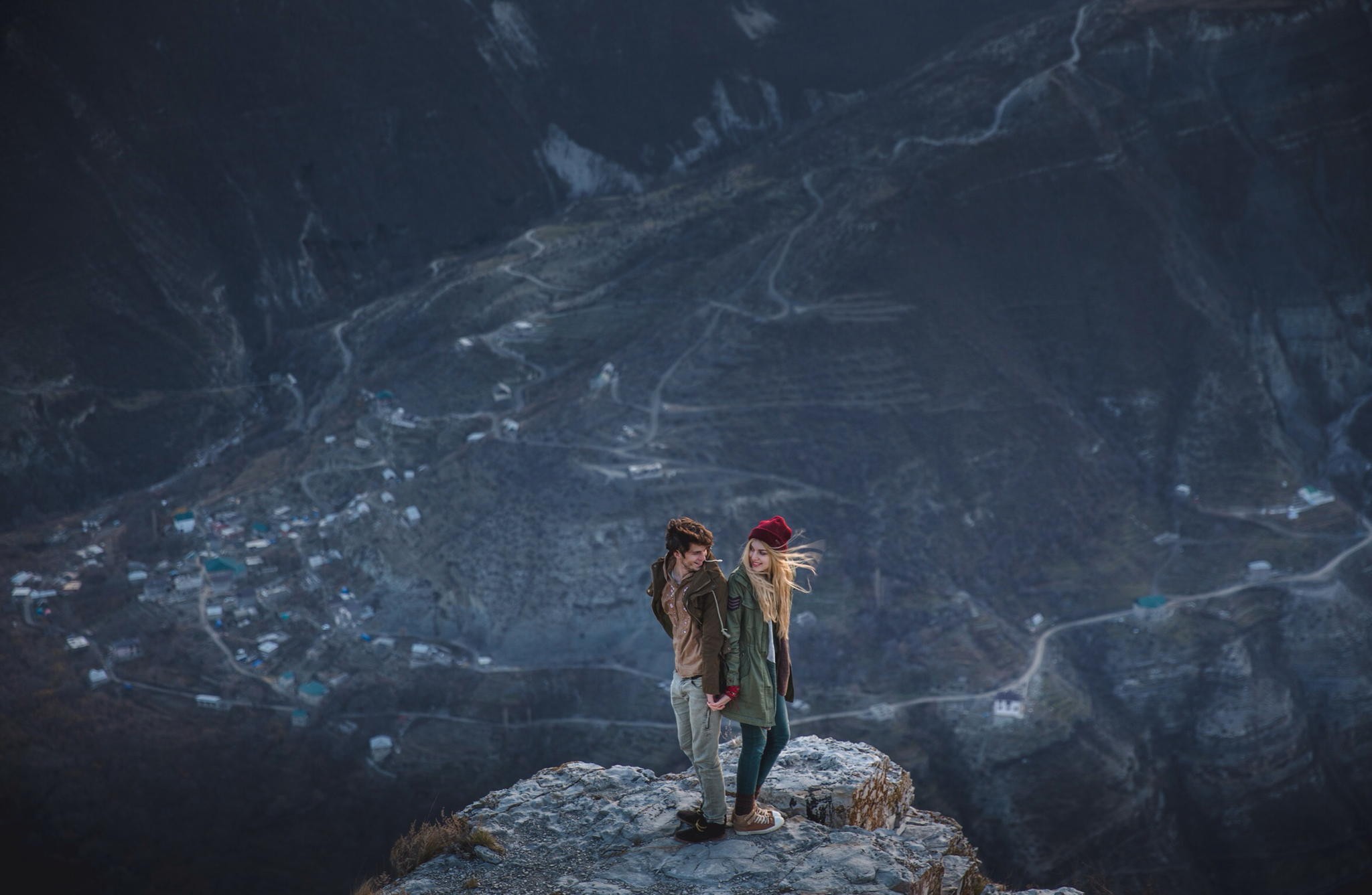 Couple standing on a cliff overlooking a mountain village HD Wallpaper