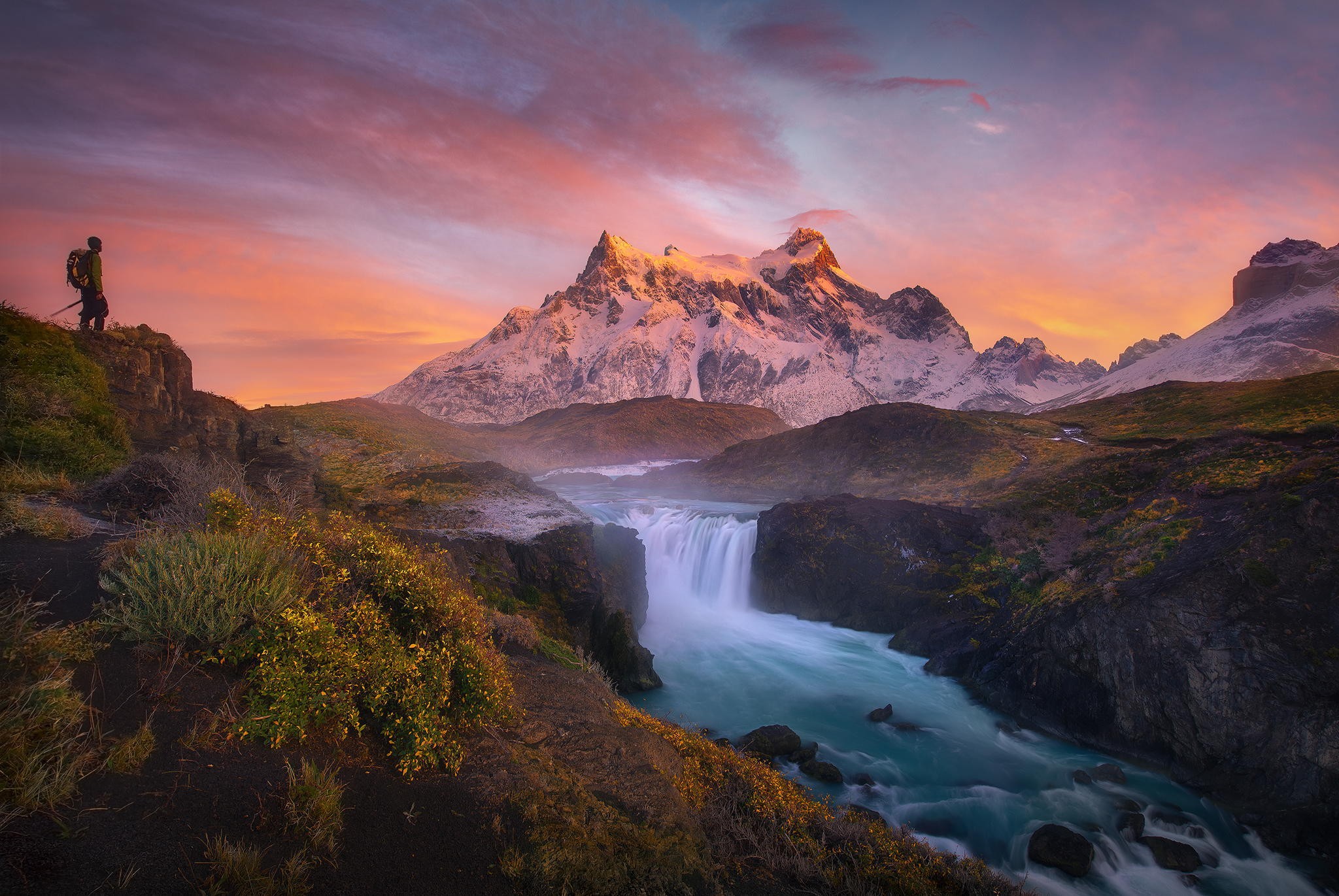 Hiker overlooking a waterfall and snowy mountains at sunset HD Wallpaper