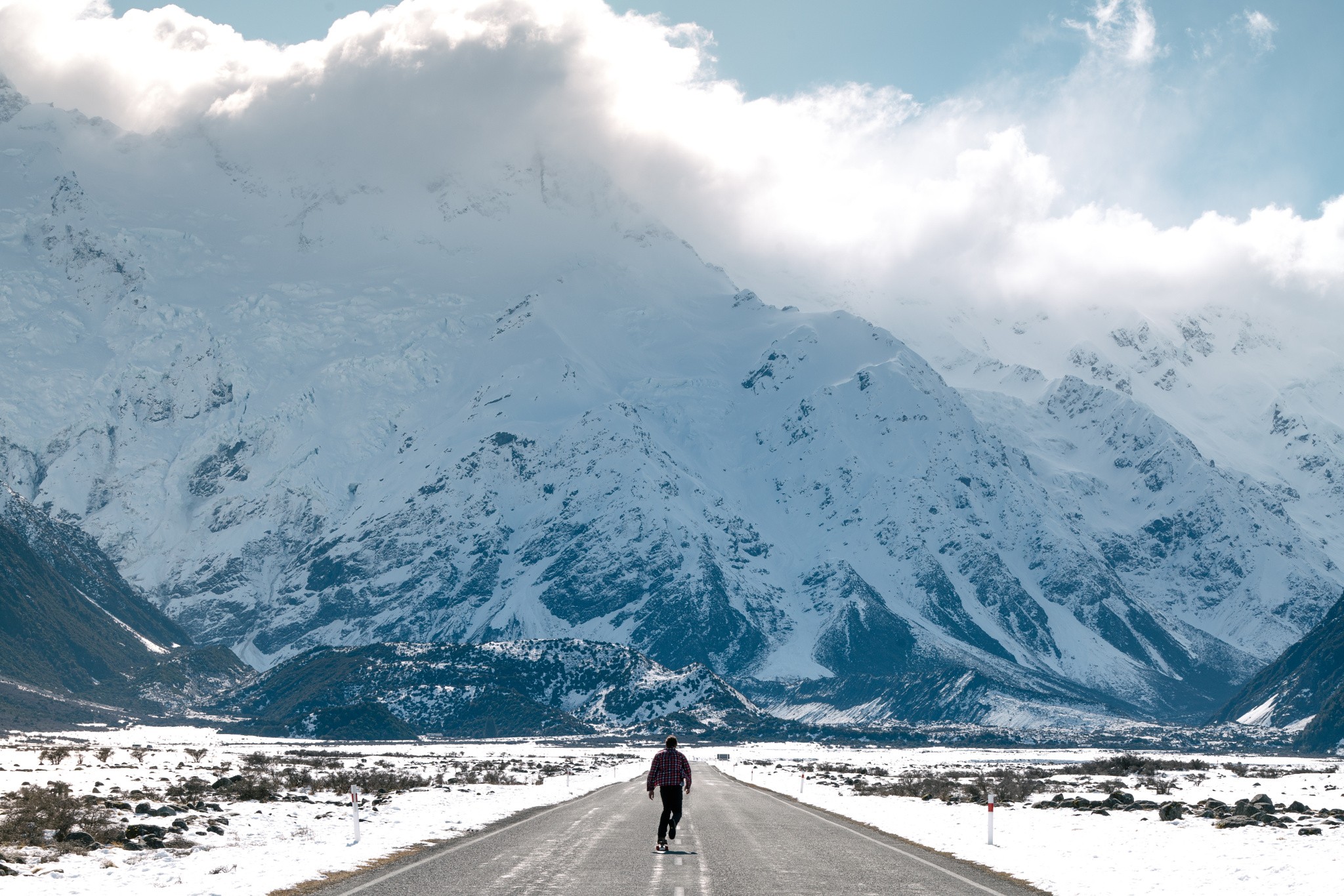 Person skateboarding on a road with snowy mountains HD Wallpaper