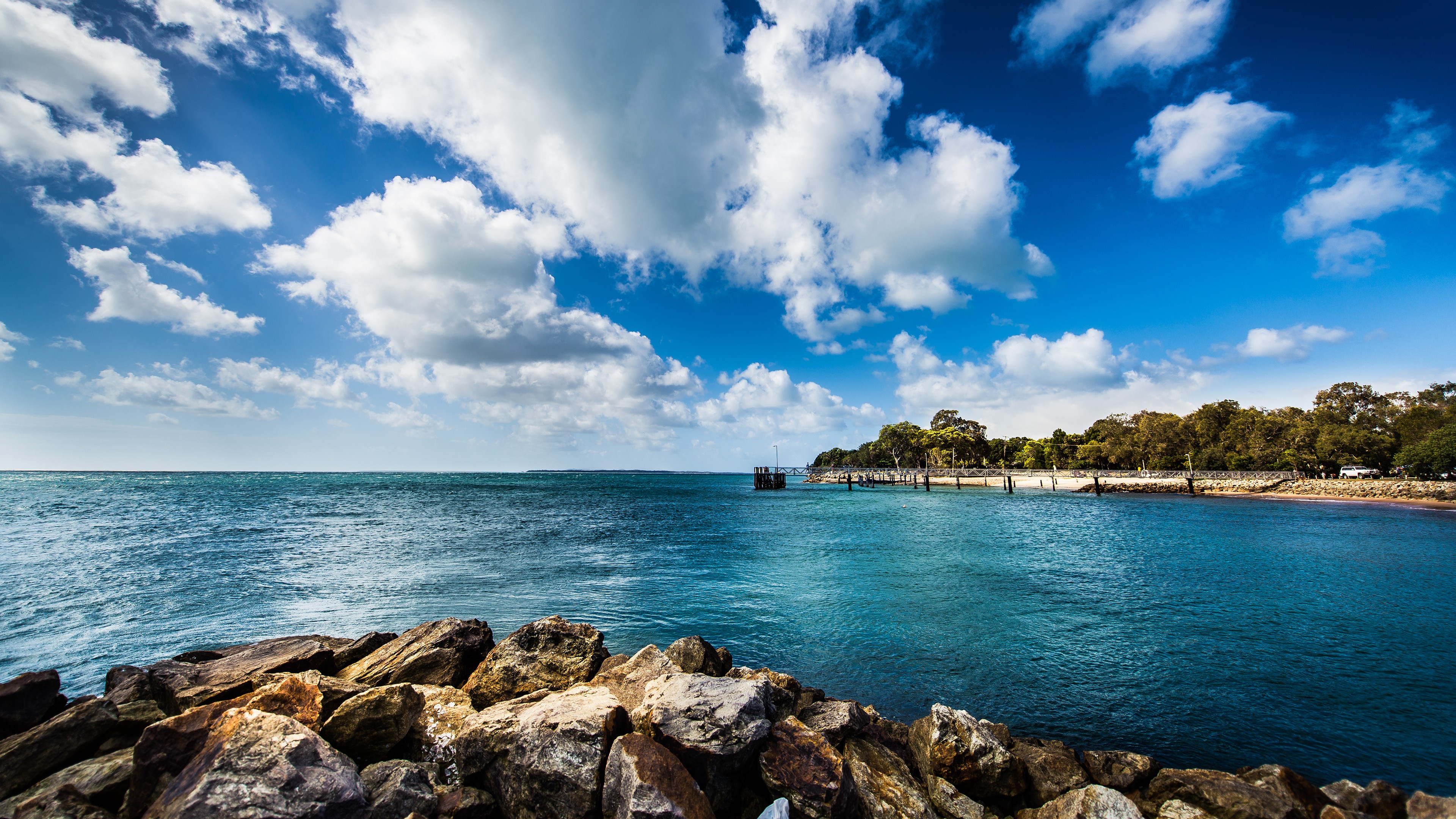 Rocky shore with a pier and trees under a cloudy sky HD Wallpaper