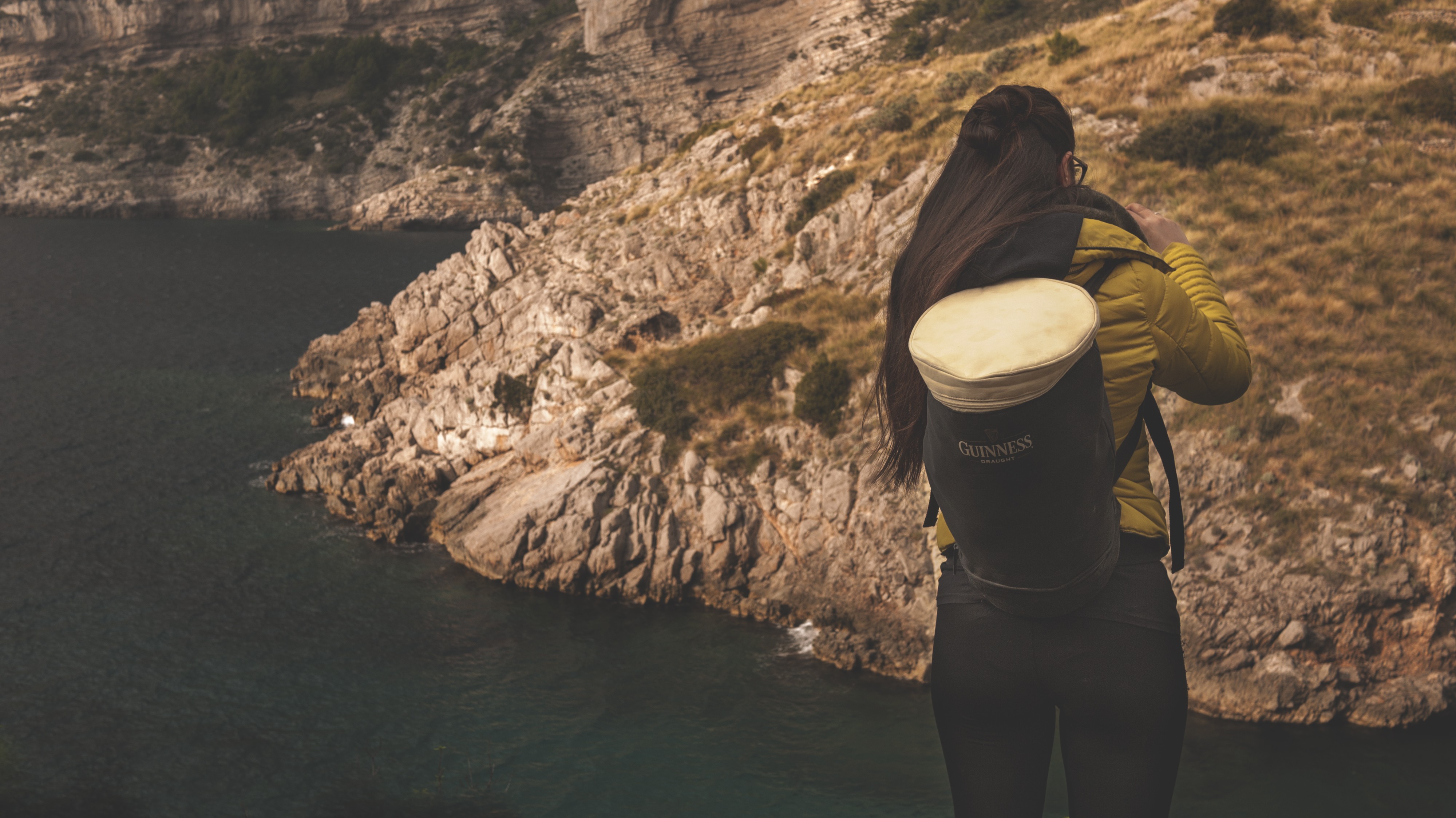 Woman with Guinness backpack on a cliff overlooking the sea HD Wallpaper