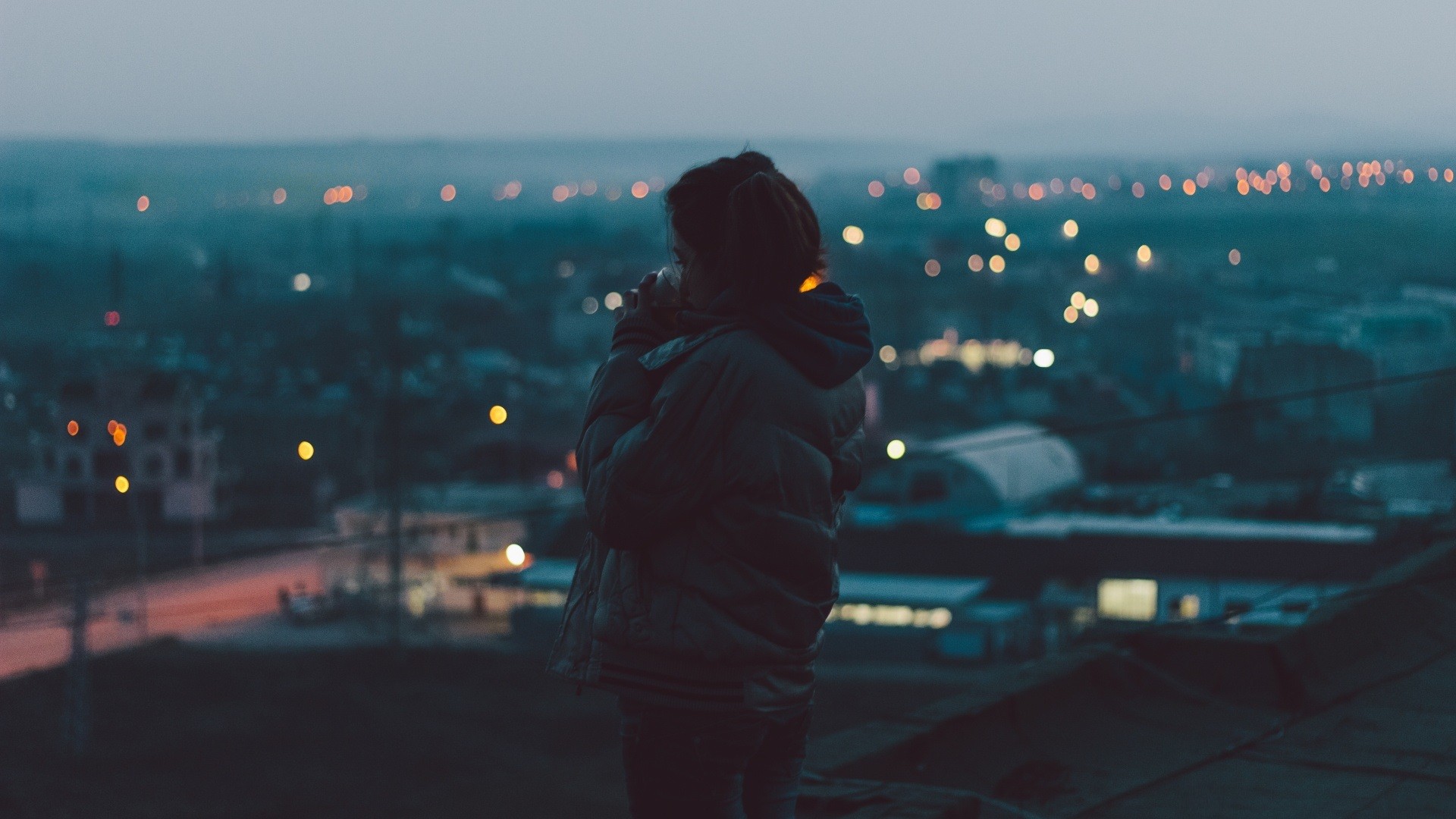Woman in winter coat looking at city lights at dusk HD Wallpaper