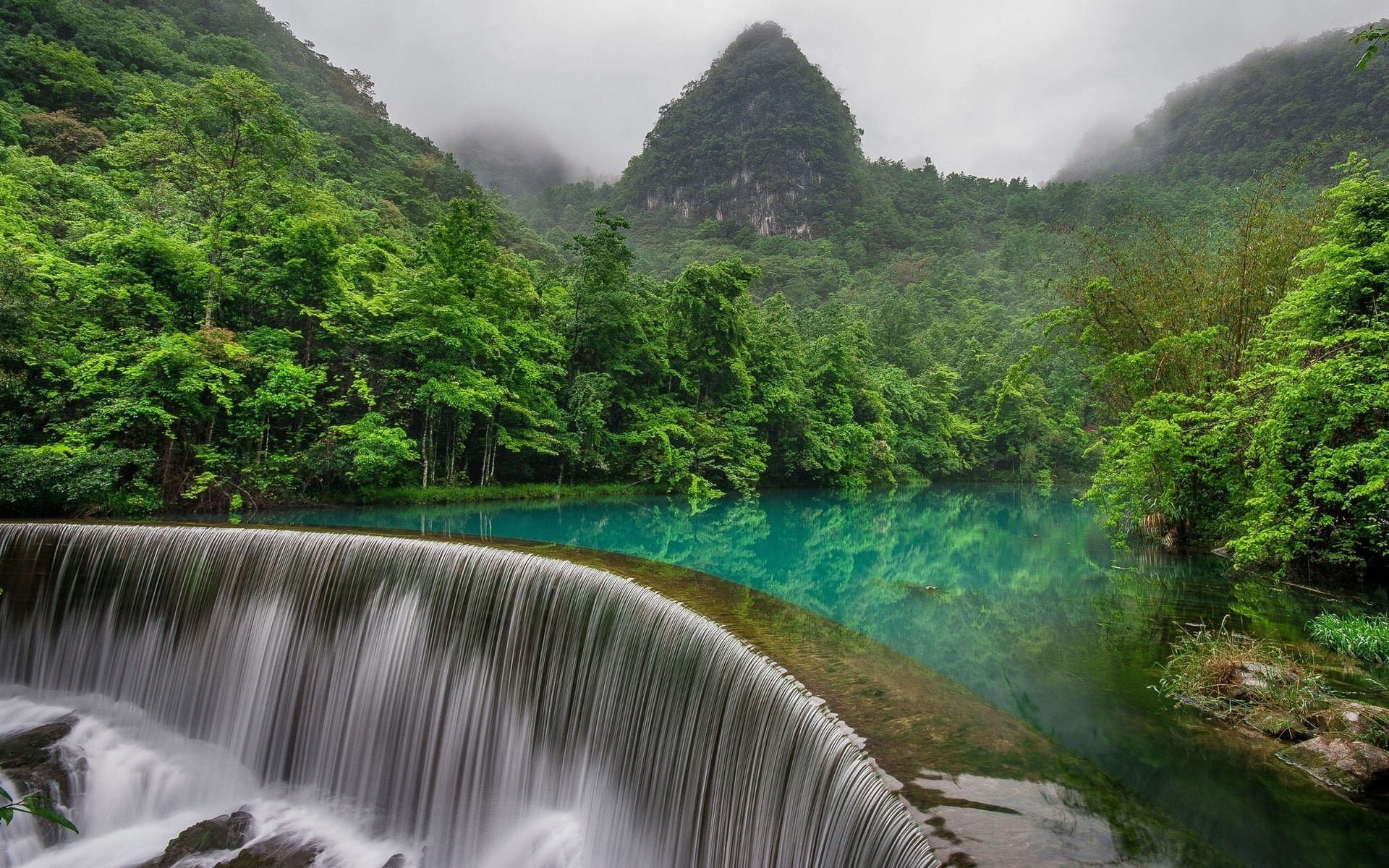 Waterfall flowing into a turquoise lake in a misty forest with mountains HD Wallpaper