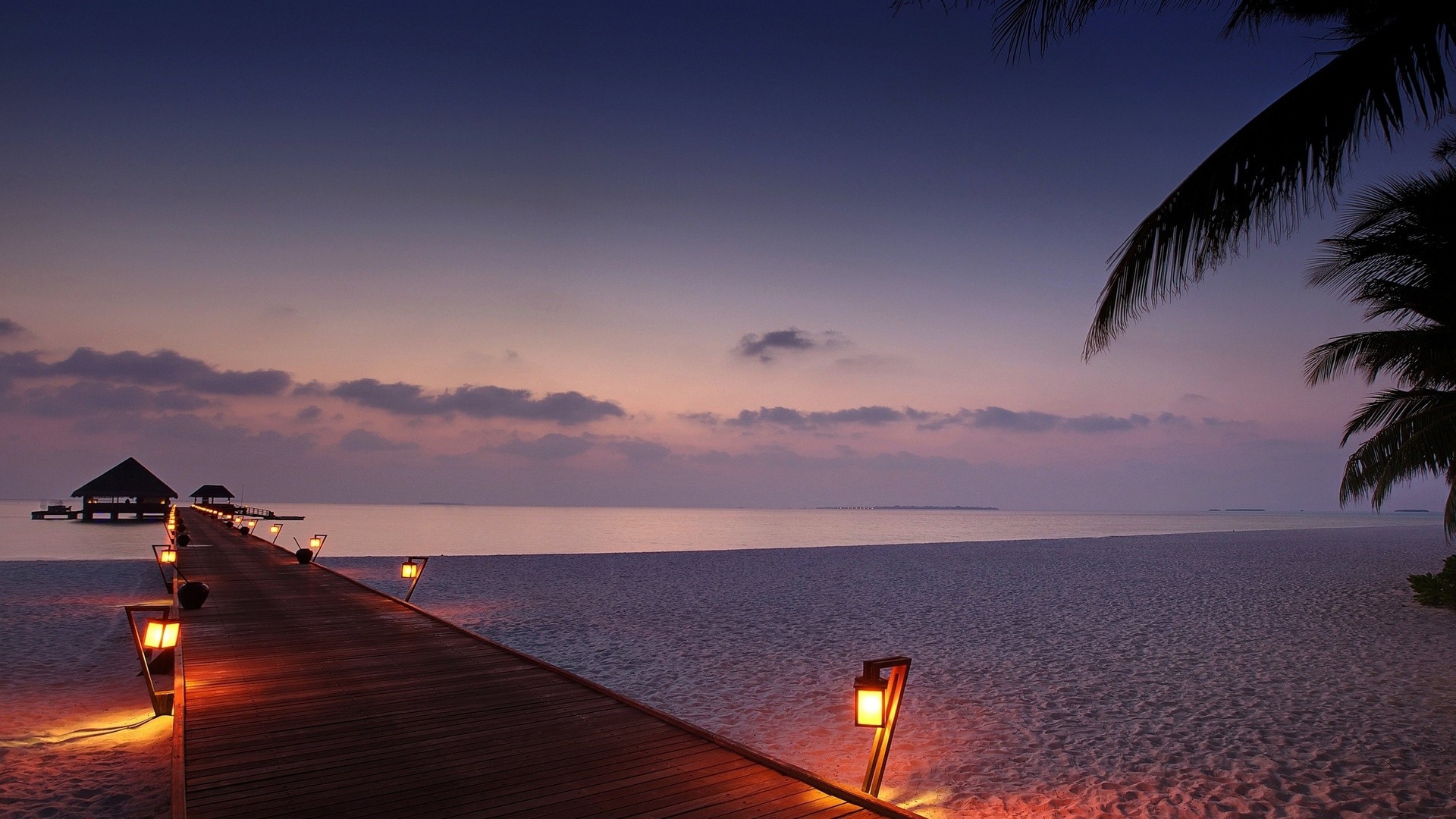 Wooden pier with lanterns leading to huts over the ocean at sunset HD Wallpaper