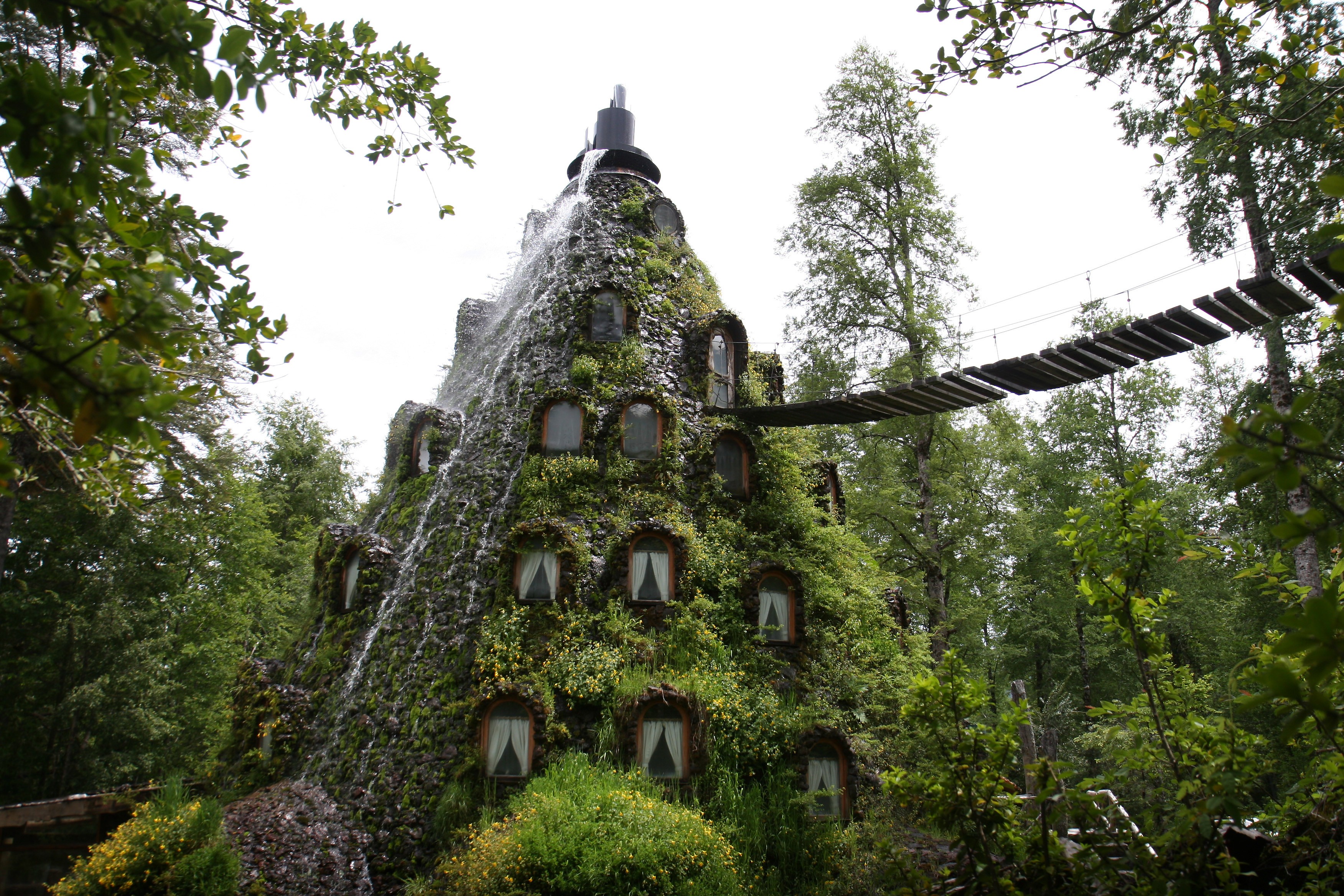 Volcanic building with waterfall and suspension bridge in forest HD Wallpaper