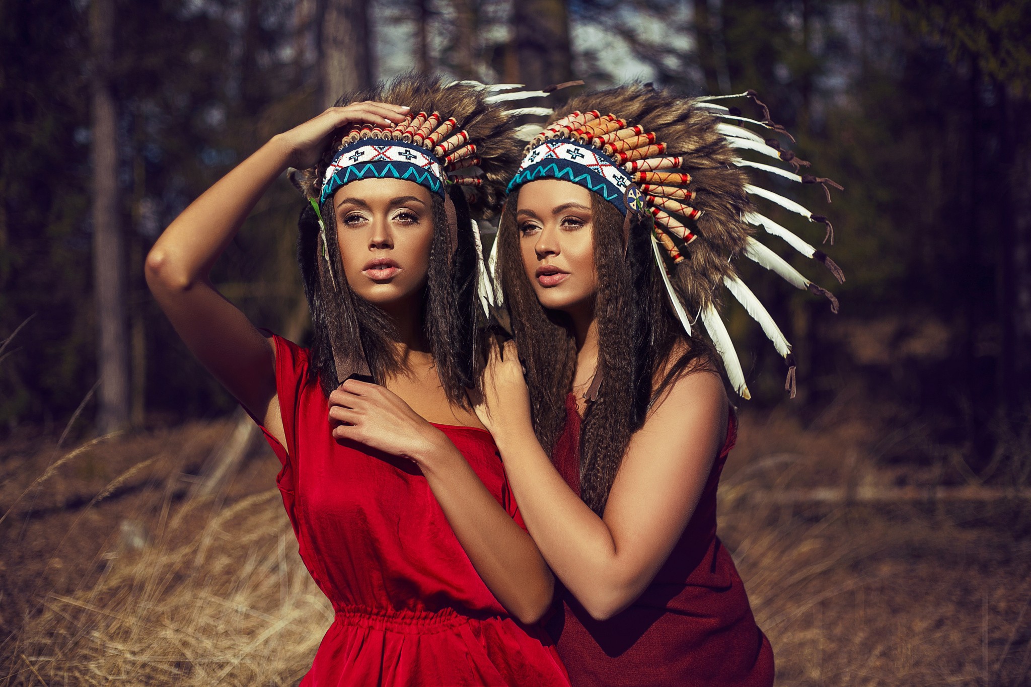 Two women wearing Native American headdresses in a forest HD Wallpaper