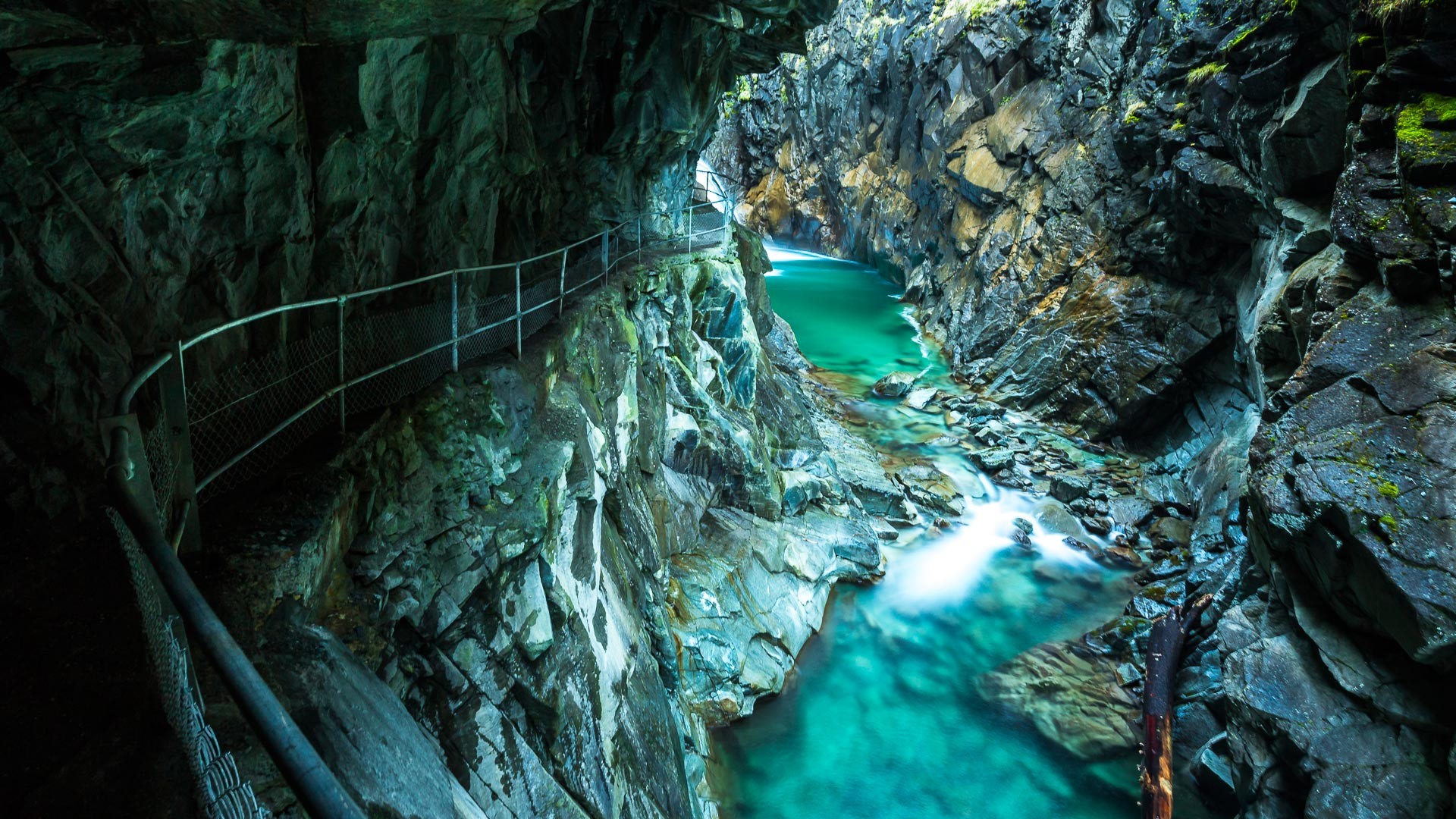 A turquoise river flows through a rocky gorge with a walkway HD Wallpaper