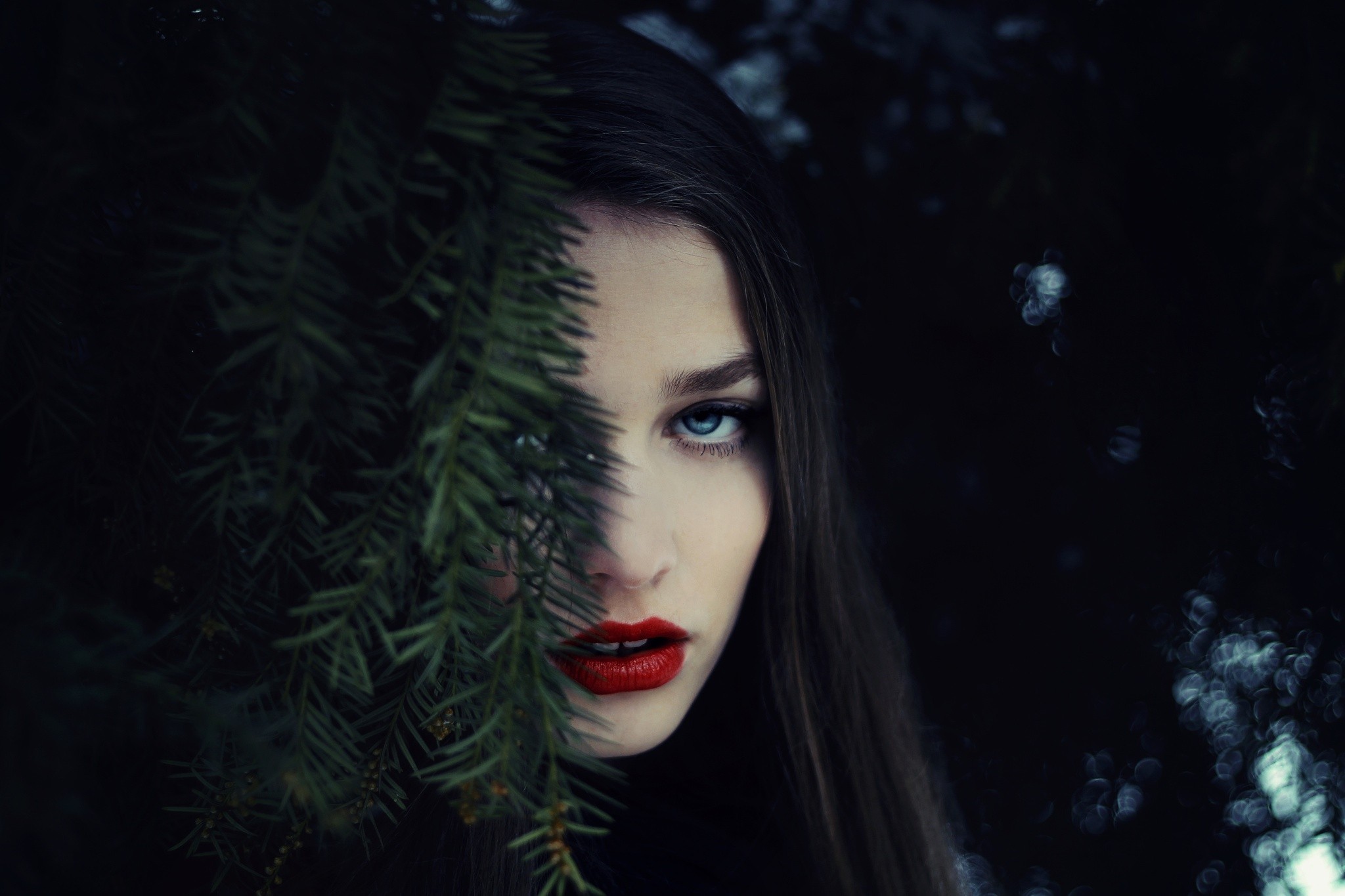 Woman with blue eyes and red lipstick behind pine branches HD Wallpaper
